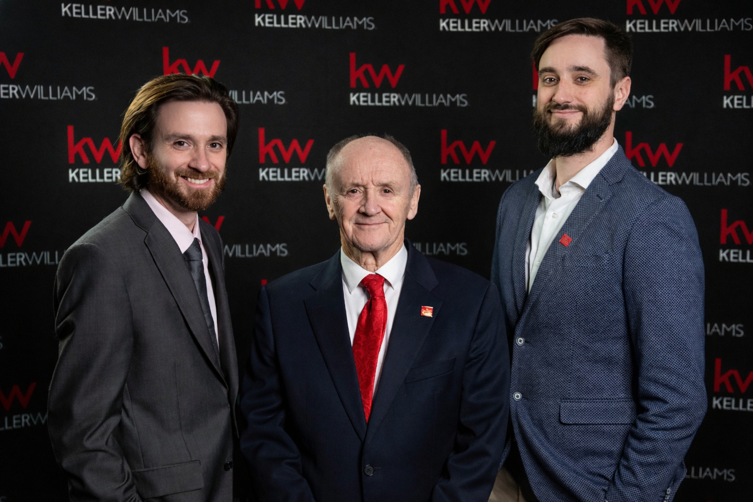 Three men in suits standing in front of a black backdrop with red and white 'Keller Williams' logos, smiling at the camera.