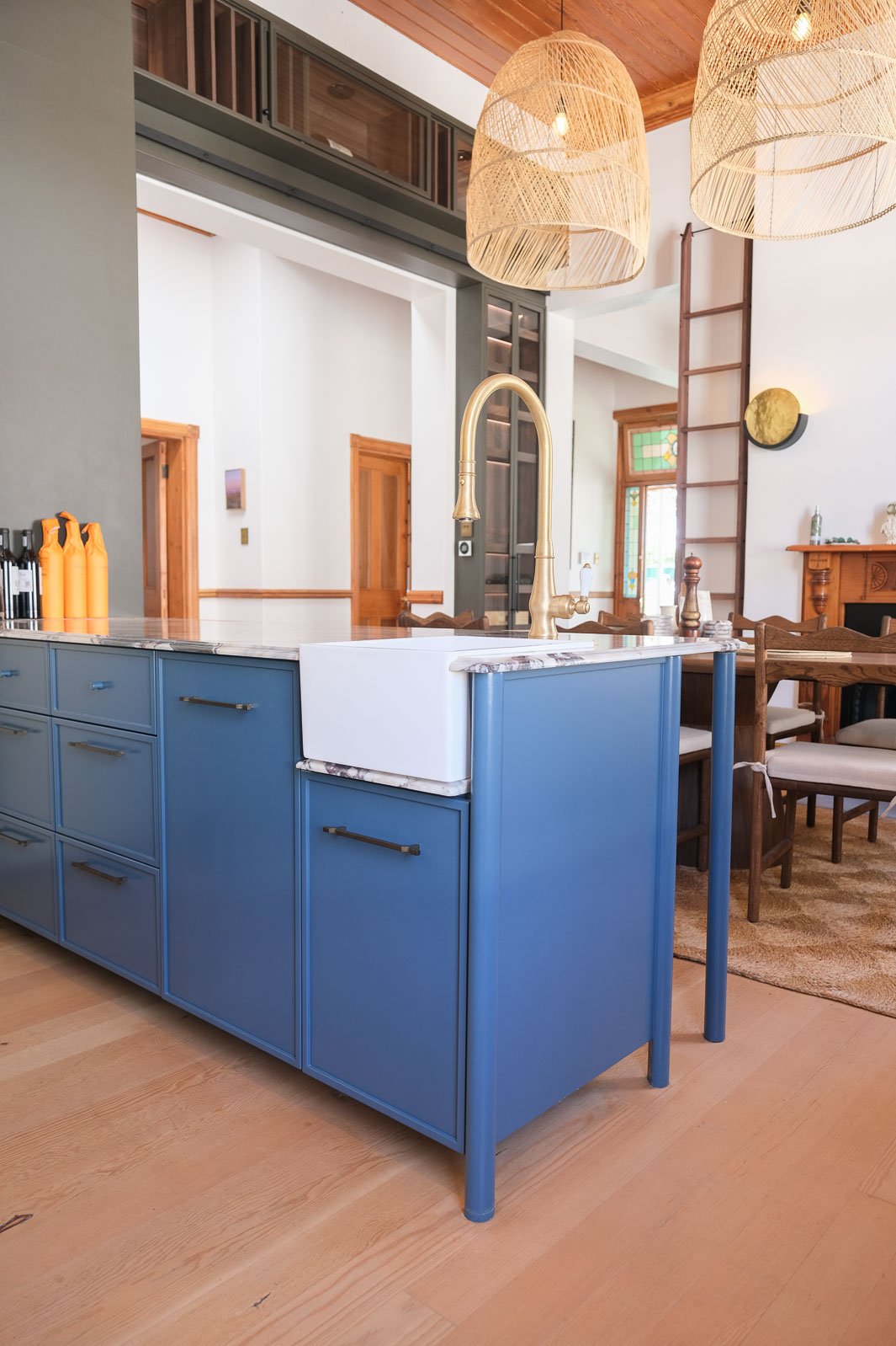 Willus Bosch, Renovation, Interior Design, Cape Town, Stellenbosch, South Africa. A modern kitchen with a blue island and a white farmhouse sink with a brass faucet. Pendant lights hang from a wood-paneled ceiling.