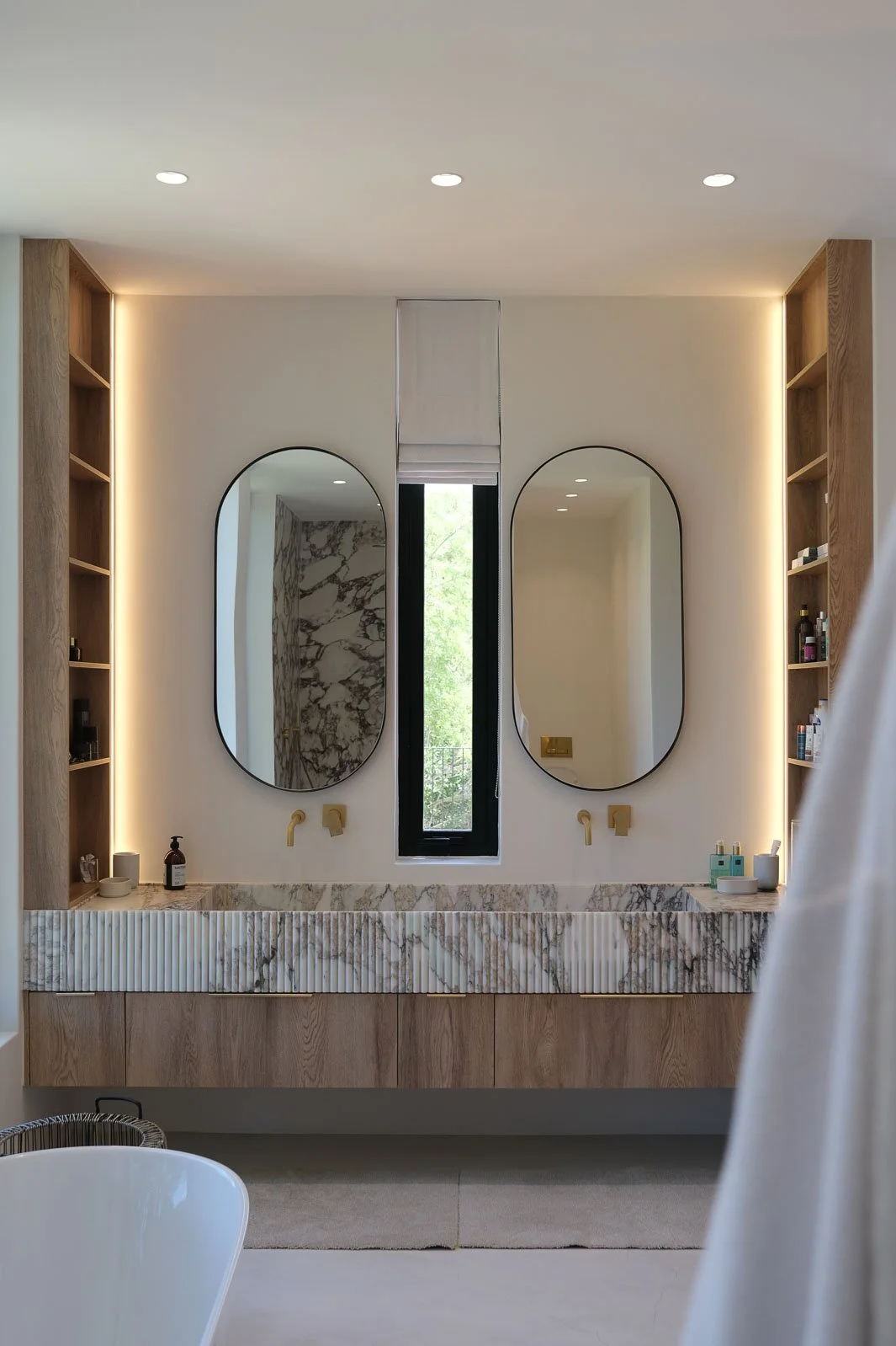 Willus Bosch, Renovation, Interior Design, Cape Town, Stellenbosch. Modern bathroom with double oval mirrors above a marble countertop, wooden cabinetry, and open shelves with toiletries, natural light from a window, and warm ceiling lighting.