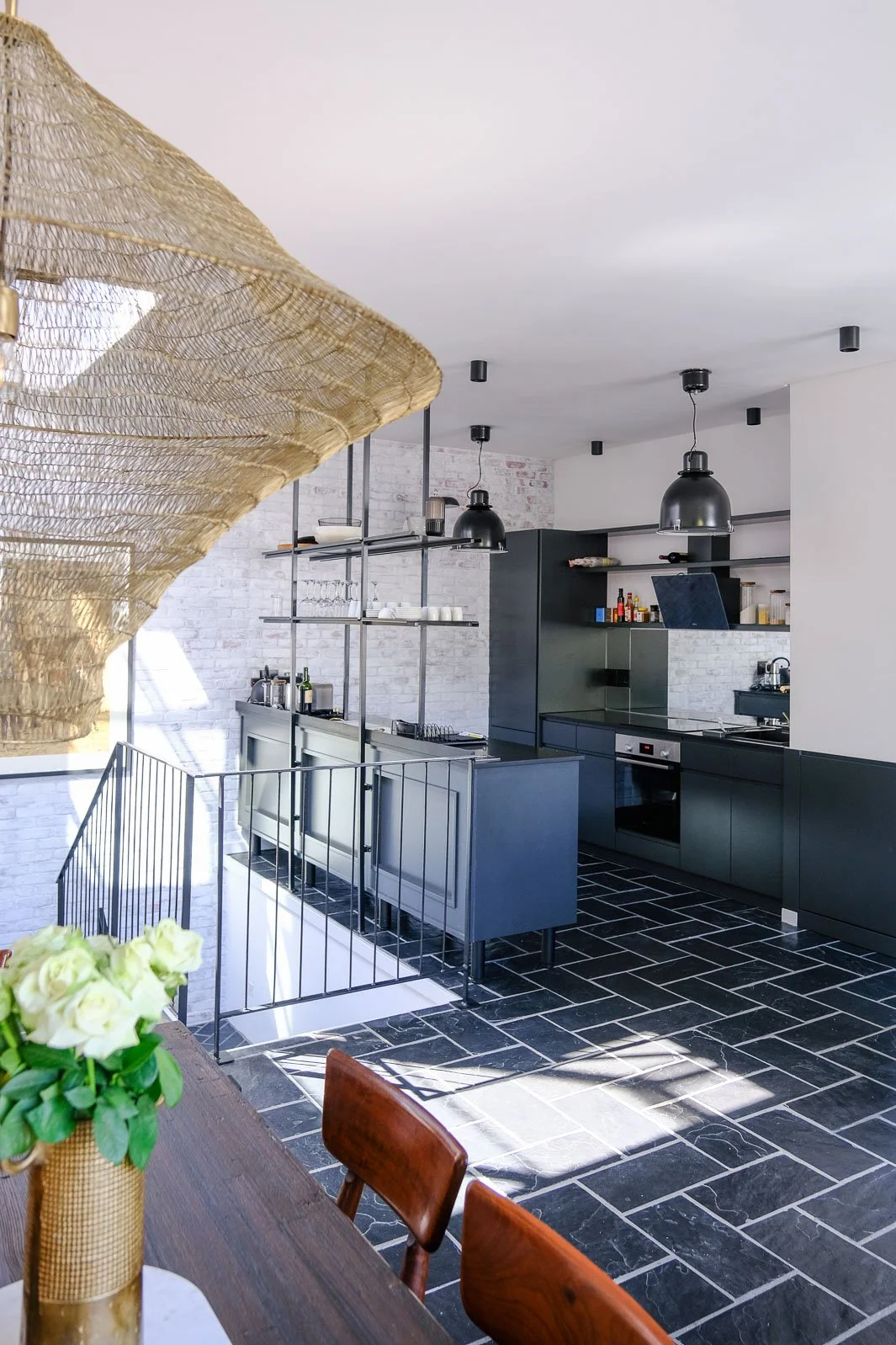 Willus Bosch, Renovation, Interior Design. Modern kitchen with black cabinets, open shelves, brick wall, black pendant lights, and black tiled floor. There is a staircase with a railing and a table with a vase of white flowers in the foreground.