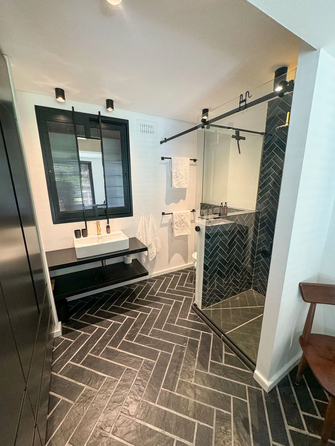 Willus Bosch, Renovation. Modern bathroom with black and white color scheme, featuring a black-framed window, a black vanity with a white sink, towel bars with towels, a glass shower with black herringbone tile, and black ceiling spotlights.