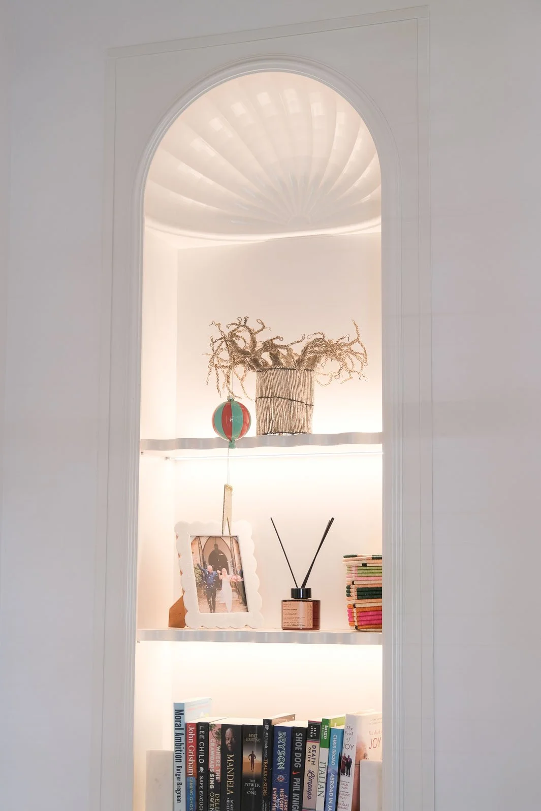 Decorative white shelving unit built into a wall with three shelves, holding a framed photograph, books, a reed diffuser, a colorful hanging ornament, a wire basket, and a dried plant arrangement on the top shelf, with a semi-circular ceiling design above.