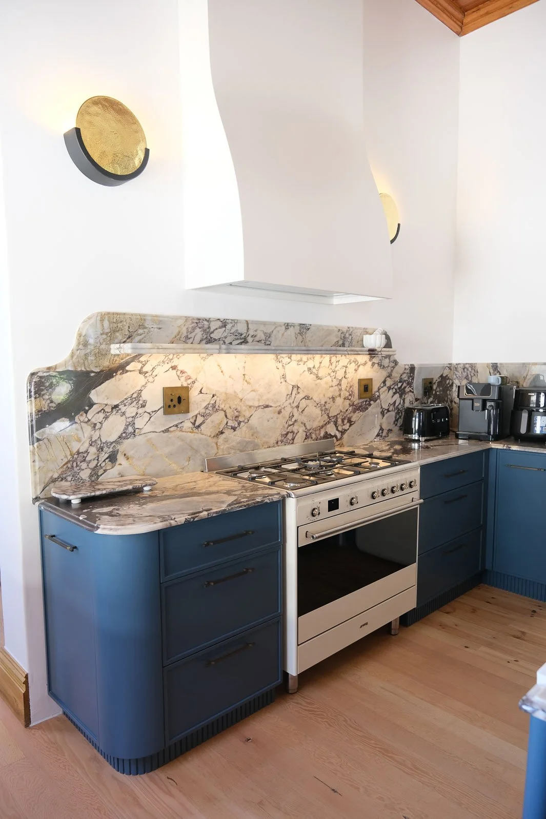 Willus Bosch, Renovation.  A modern kitchen with blue cabinets, a marble backsplash, and a white stove. On the countertop, there are small appliances, including a coffee maker and a toaster. The wall has decorative gold and black wall lights.