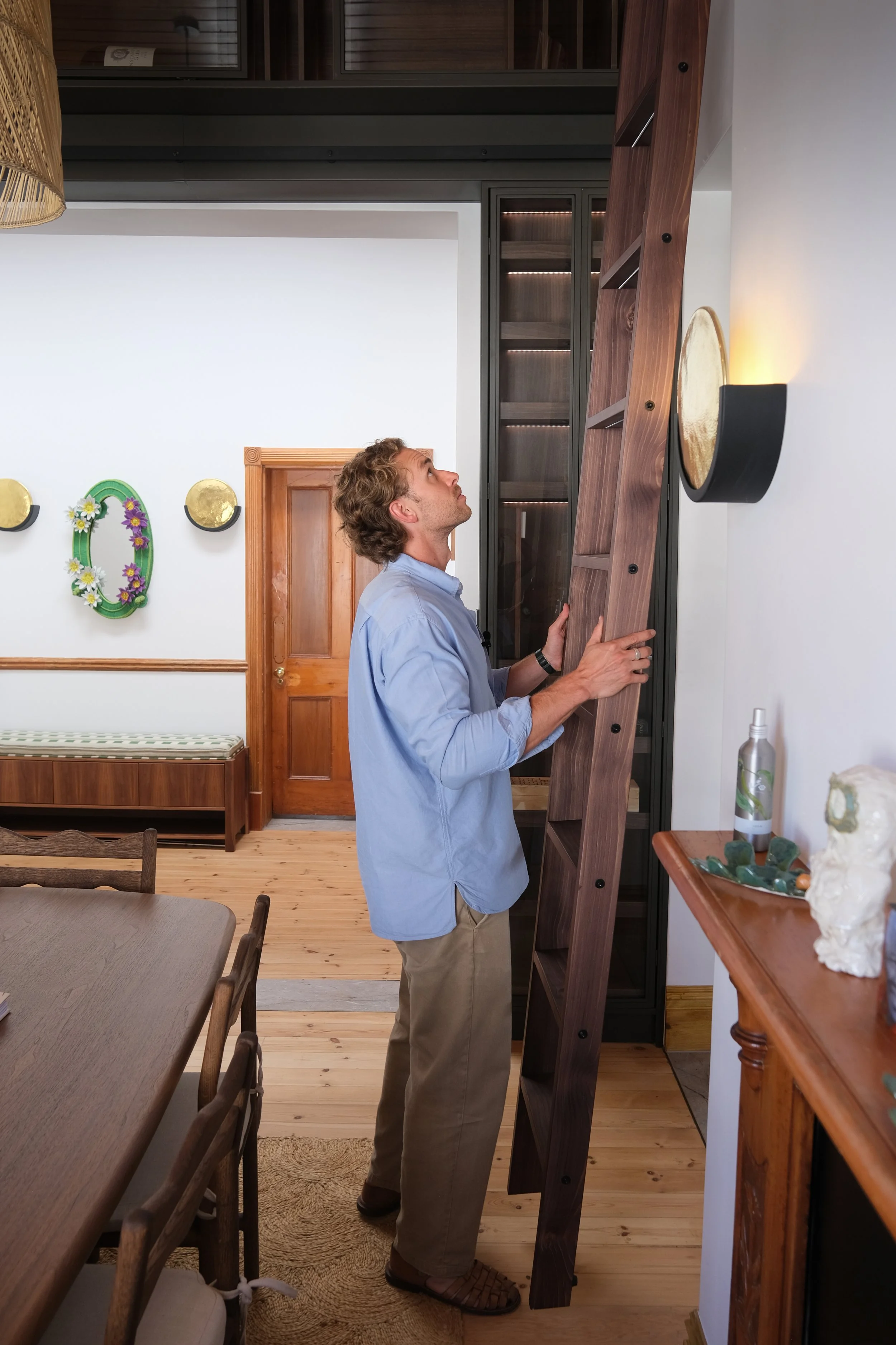 Willus Bosch, Renovation, Interior Design, Cape Town, Stellenbosch, South Africa. A man with curly hair wearing a light blue shirt and khaki pants looking up at a tall wooden shelf or furniture piece in a living room.