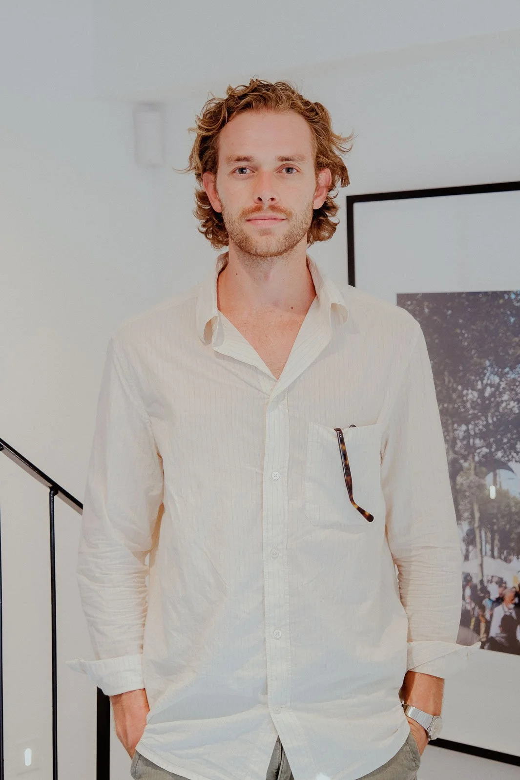 Willus Bosch, Renovation, Interior Design, Cape Town, Stellenbosch, South Africa. A young man with wavy, shoulder-length hair and a light beard, wearing a cream-colored button-up shirt, standing indoors with hands in pockets.