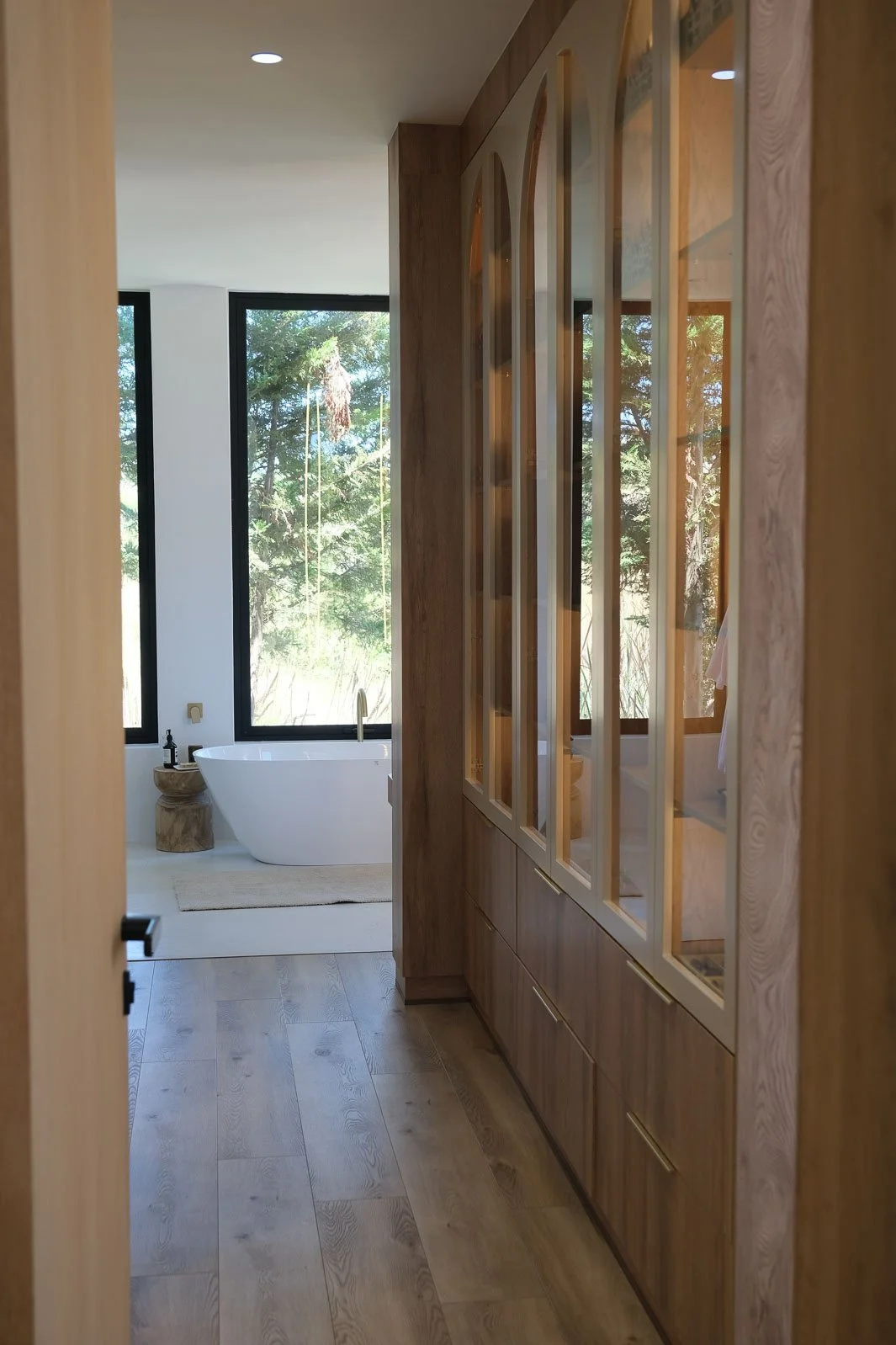 Willus Bosch, Renovation, Interior Design, Cape Town, Stellenbosch, South Africa. View of a modern bathroom with a freestanding bathtub near large windows showing trees outside, a wooden side table, and wooden cabinets with glass doors.