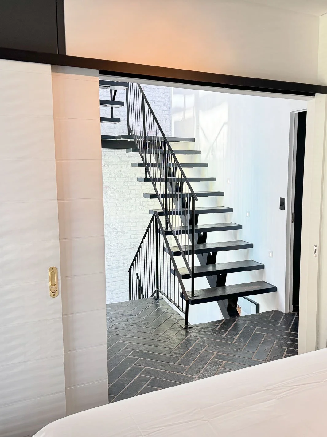 Willus Bosch, Renovation, Interior Design, Cape Town. Modern black metal staircase with open risers and a black railing, leading to an upper floor in a bright, contemporary home with white and gray walls and dark herringbone tile flooring.
