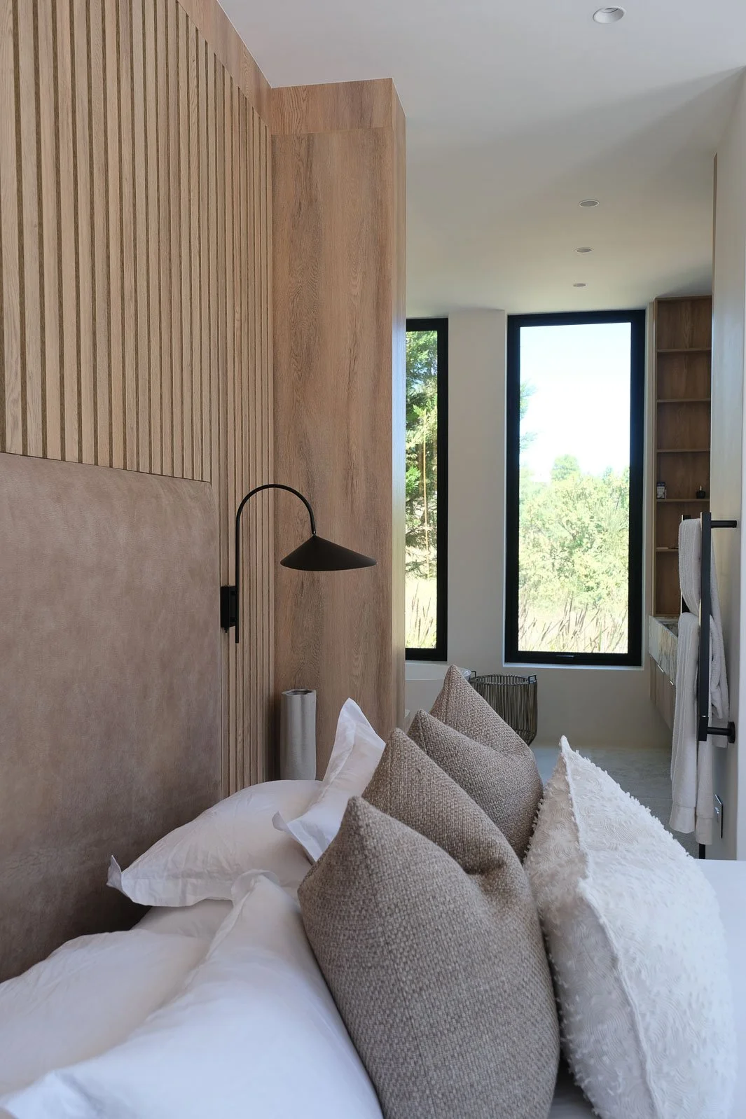 Willus Bosch. A cozy bedroom with a bed that has white linens and multiple beige and white pillows. There is a black wall-mounted reading lamp next to the bed. The room features wood-paneled walls and large window with a view of green trees outside.