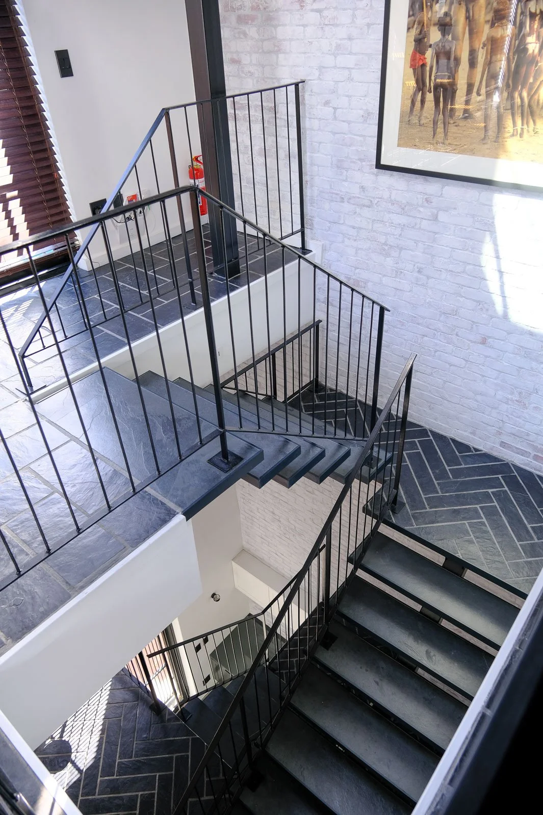 Willus Bosch, Renovation, Interior Design, Cape Town, Stellenbosch, South Africa. Interior view of a modern multi-story staircase with black railings, gray tile flooring, a white brick wall, a framed beach photograph, and a fire extinguisher.