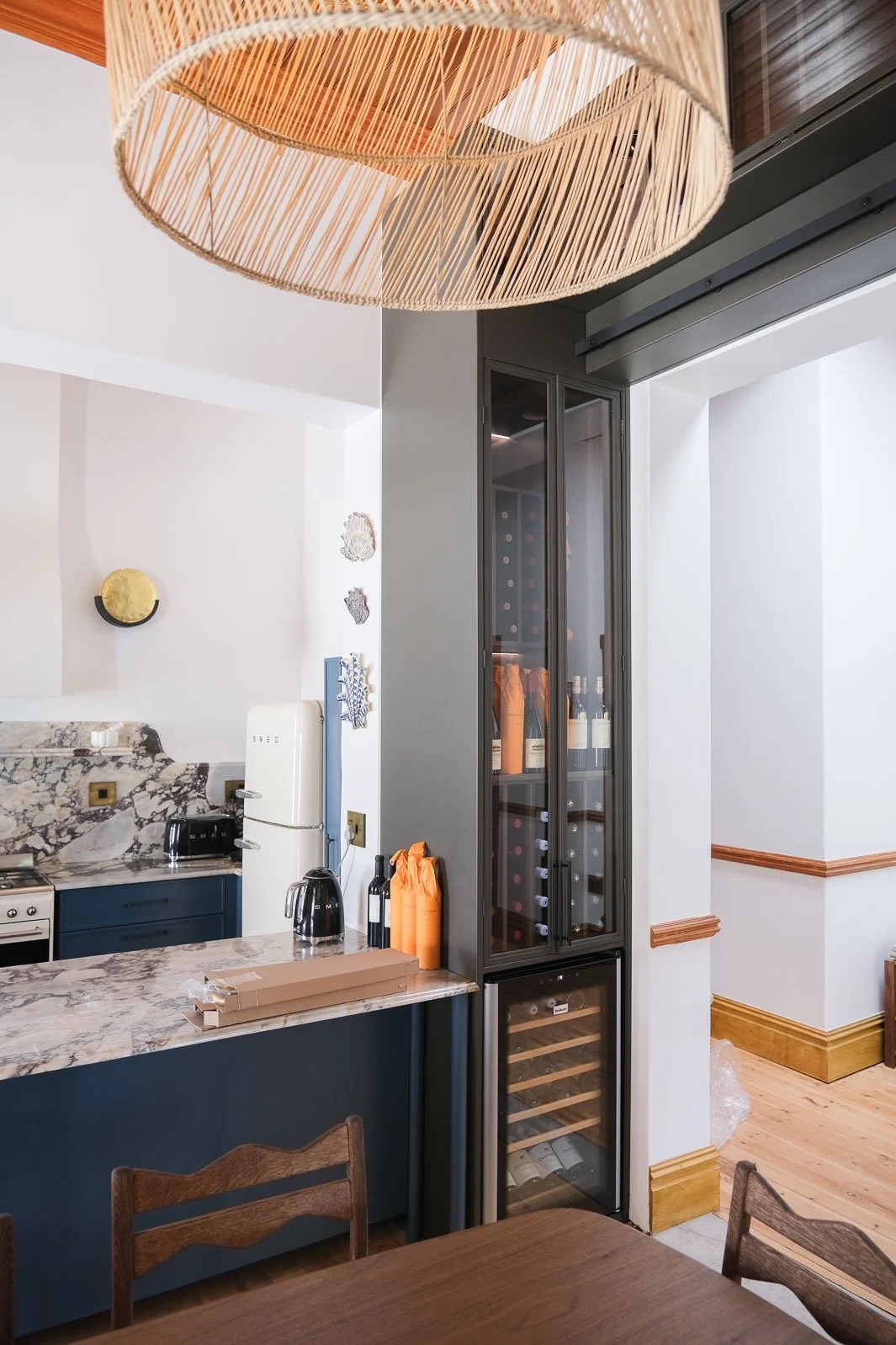 Willus Bosch, Renovation. A modern kitchen with a marble countertop, a white fridge, and a built-in wine fridge with bottles stored inside. The kitchen features dark blue cabinets, a wooden table, and a woven pendant light hanging from the ceiling.