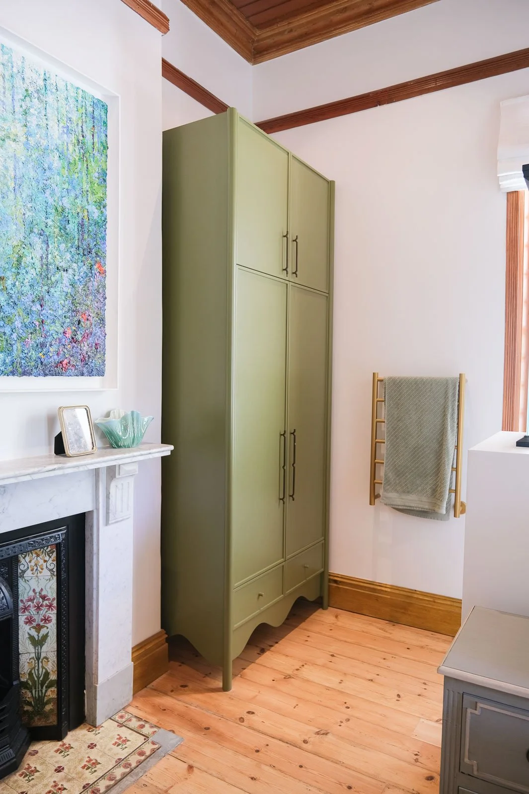 Willus Bosch, Renovation, Interior Design, Cape Town, Stellenbosch, South Africa. A corner of a room with a green storage cabinet, a fireplace with decorative tiles, a wall with artwork, a towel rack, and wooden trim on ceiling and floor.