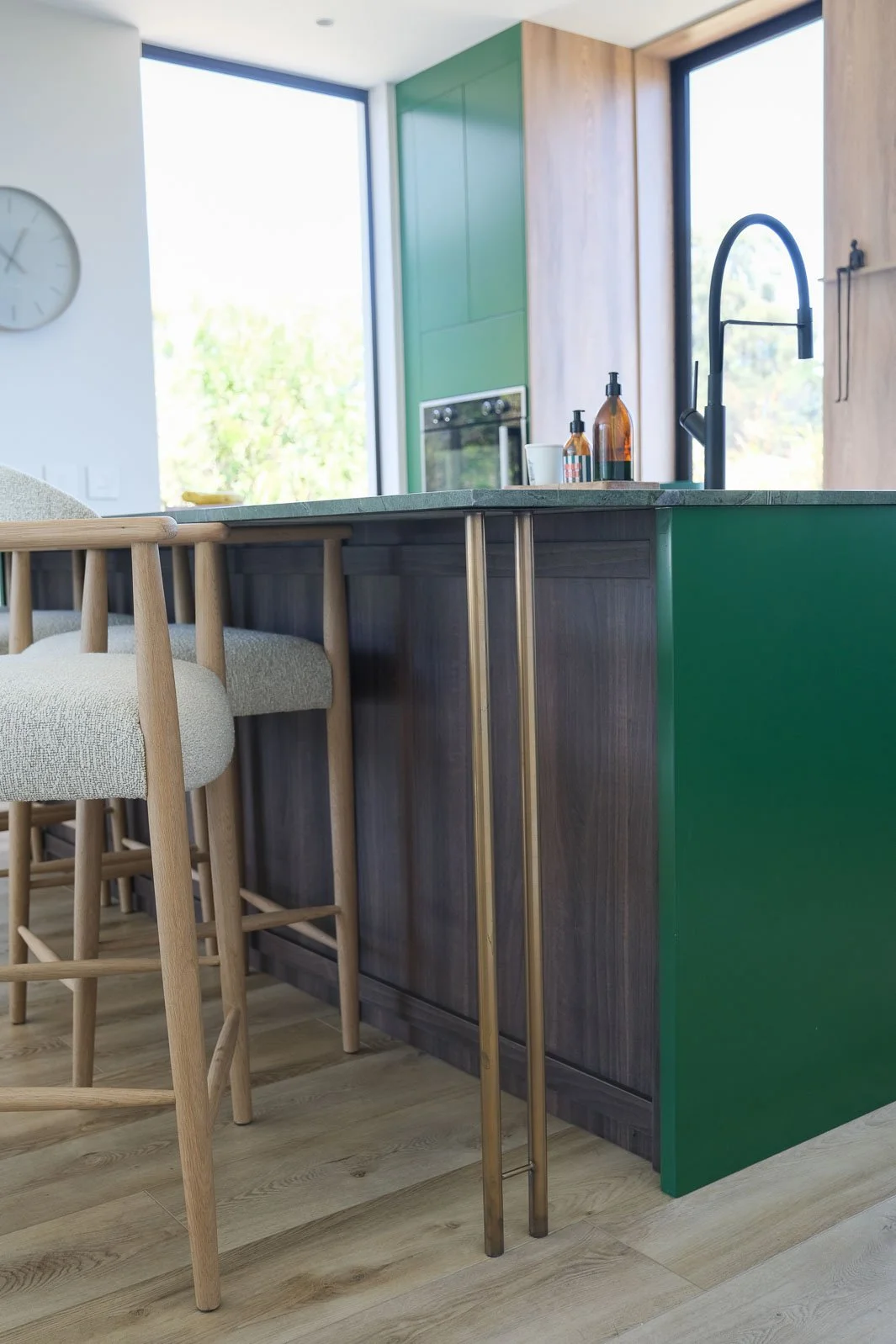 Willus Bosch, Renovation, Interior Design, Cape Town, Stellenbosch, South Africa. Close-up of kitchen island with green and wood paneling, whiskey bottles, and black faucet, with barstools nearby and large windows in the background.