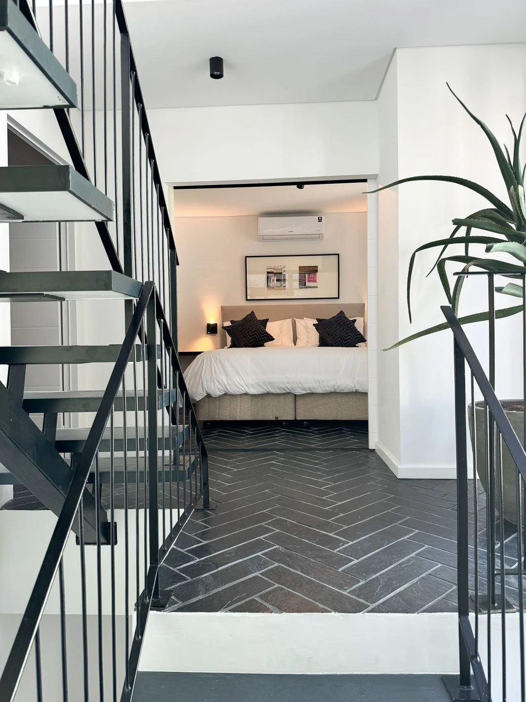 Willus Bosch, Renovation, Interior Design, Cape Town, Stellenbosch, South Africa. Modern hotel room with a bed, framed artwork, air conditioning, and a large potted plant, viewed through a staircase railing.