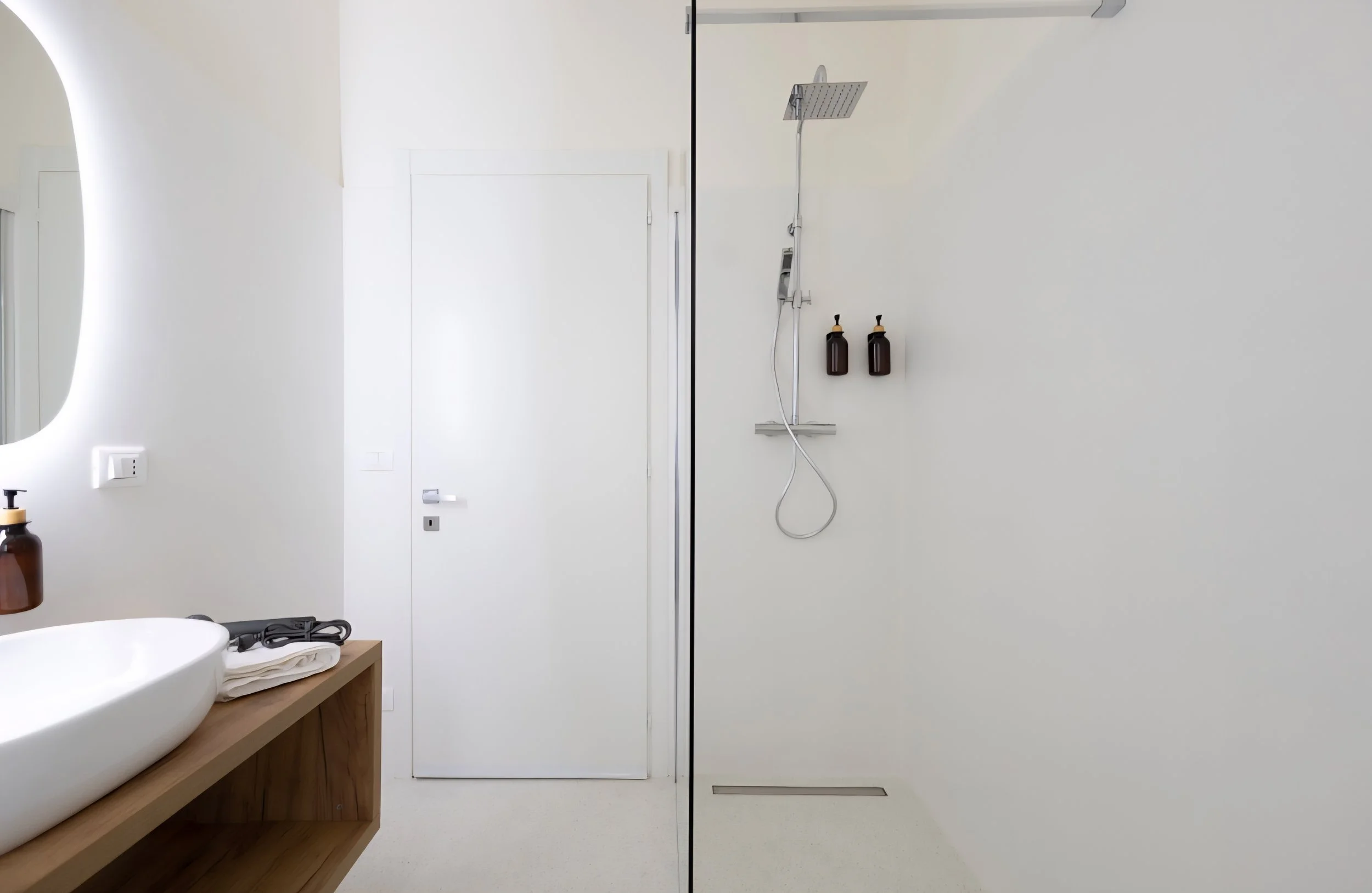 A destra, una doccia moderna con soffione e due dispenser di sapone. A sinistra, un mobile in legno con un lavabo bianco e alcuni asciugamani.