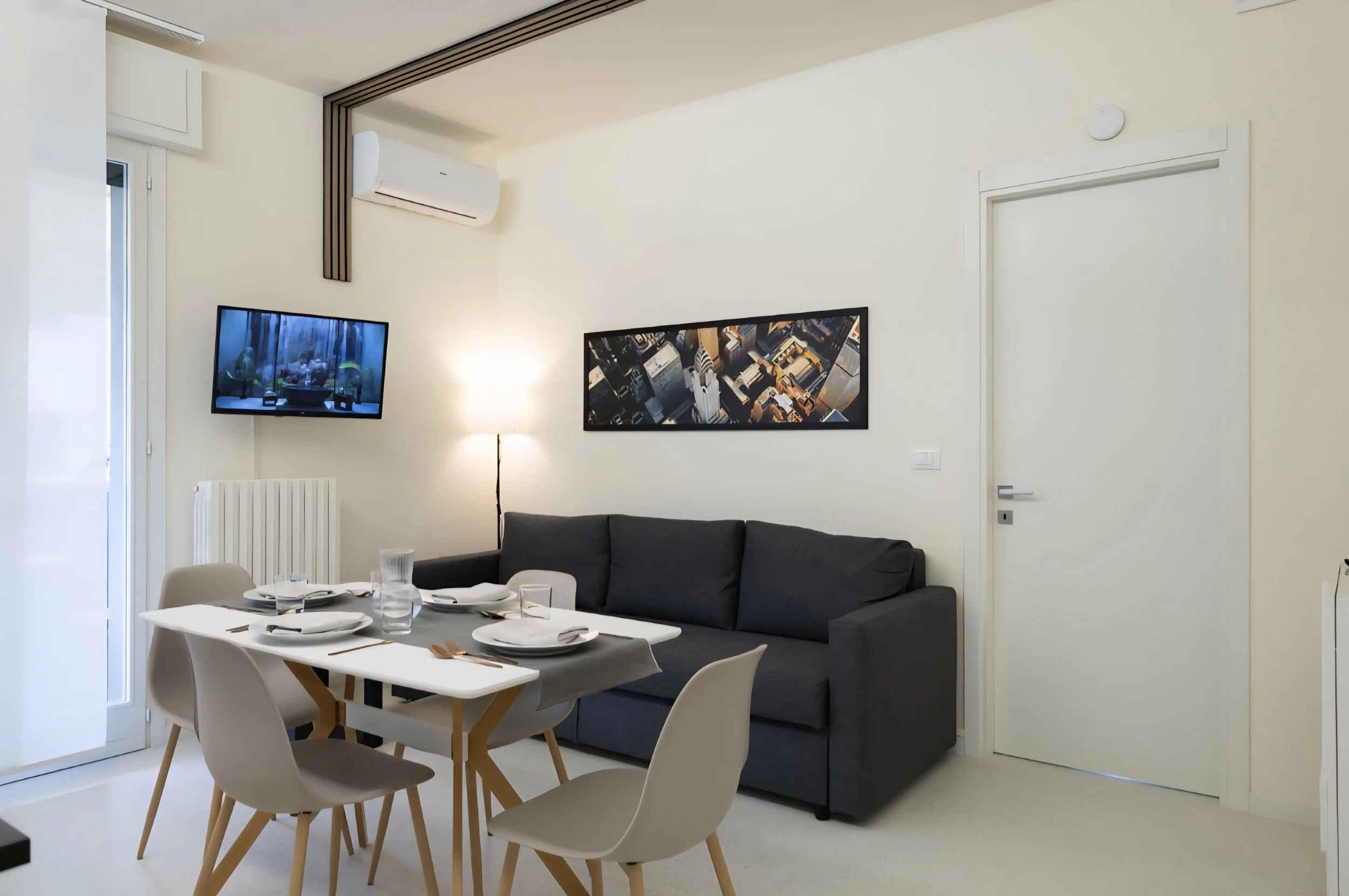Interno di un soggiorno con tavolo da pranzo apparecchiato, divano grigio, muro bianco con un quadro e un televisore, illuminazione soffusa.