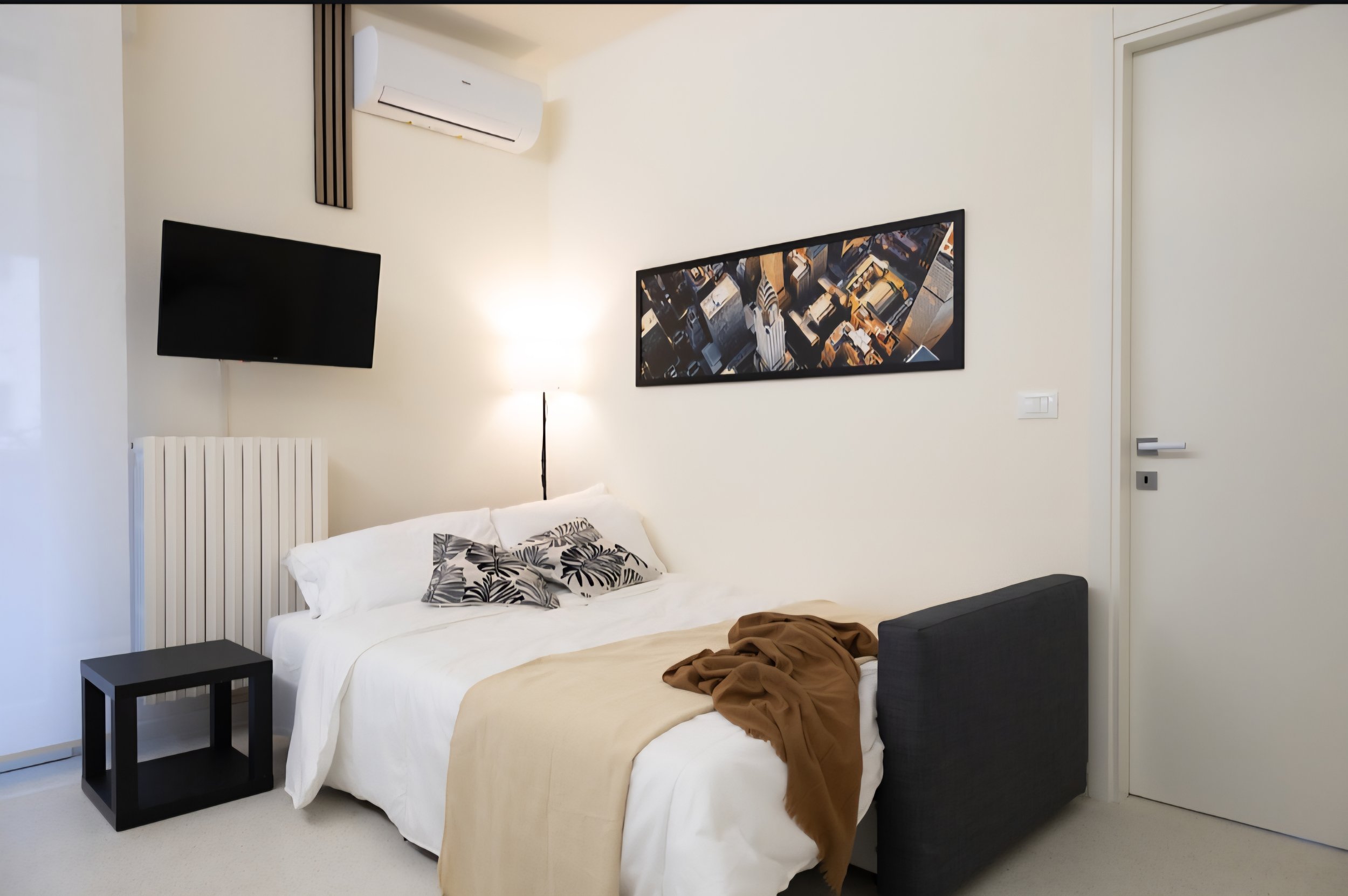 Stanza moderna e minimalista con letto, cuscini decorativi, lampada, televisore e quadro sul muro, pareti bianche e porta chiusa.