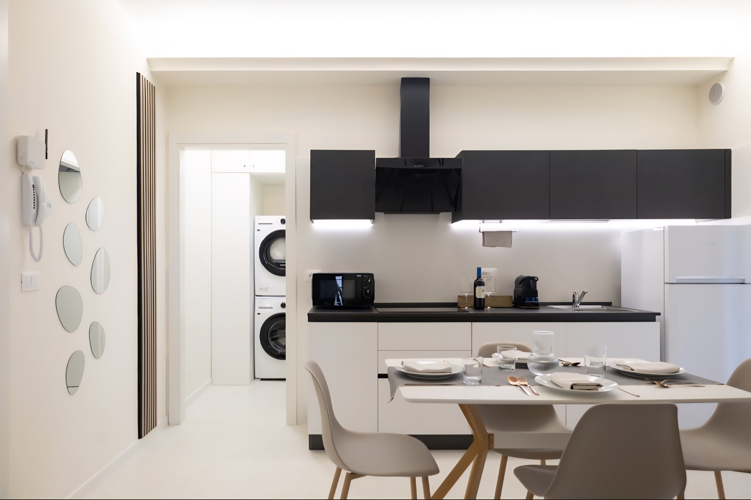 Cucina moderna bianca con mobili neri, tavolo apparecchiato con piatti e posate, in background lavatrice e asciugatrice, ripiani e elettrodomestici