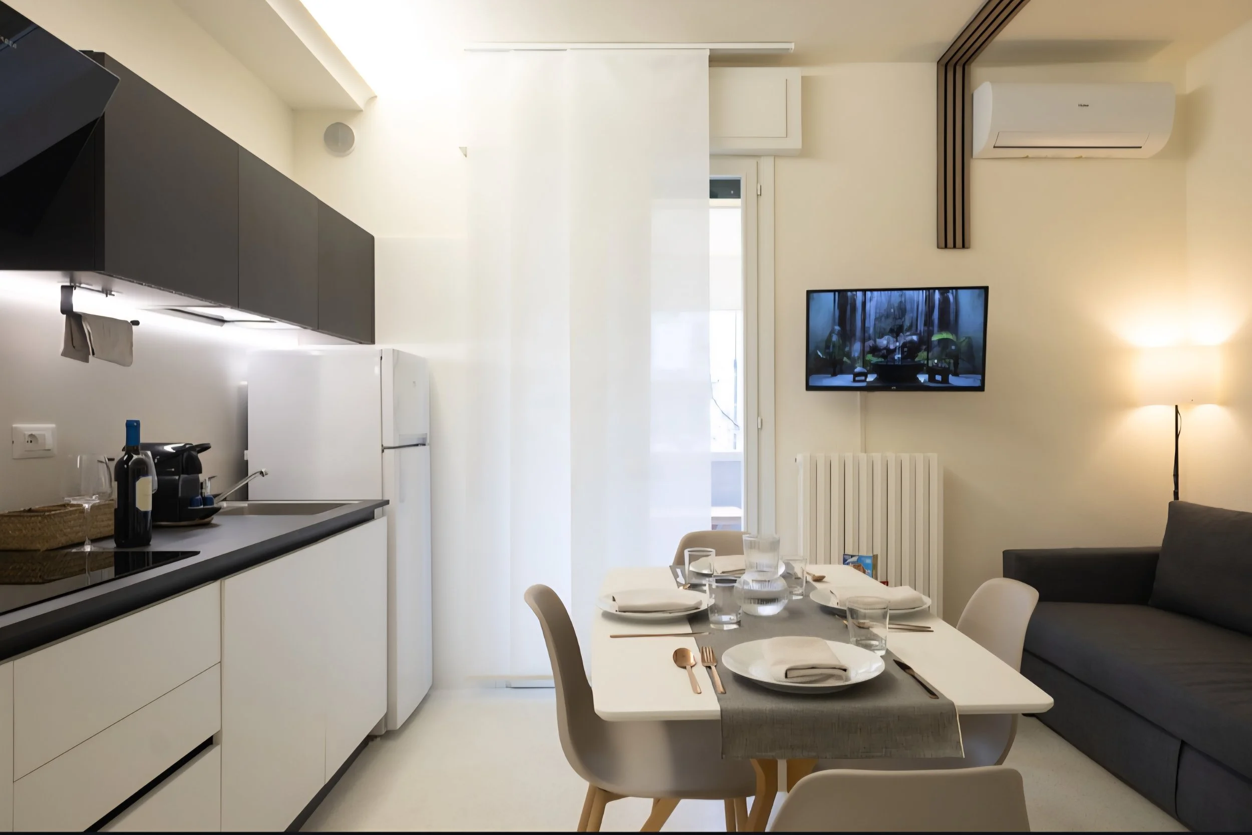 Interno di un soggiorno moderno con cucina, tavolo apparecchiato per quattro persone, divano grigio, televisionе, lampada da terra e aria condizionata