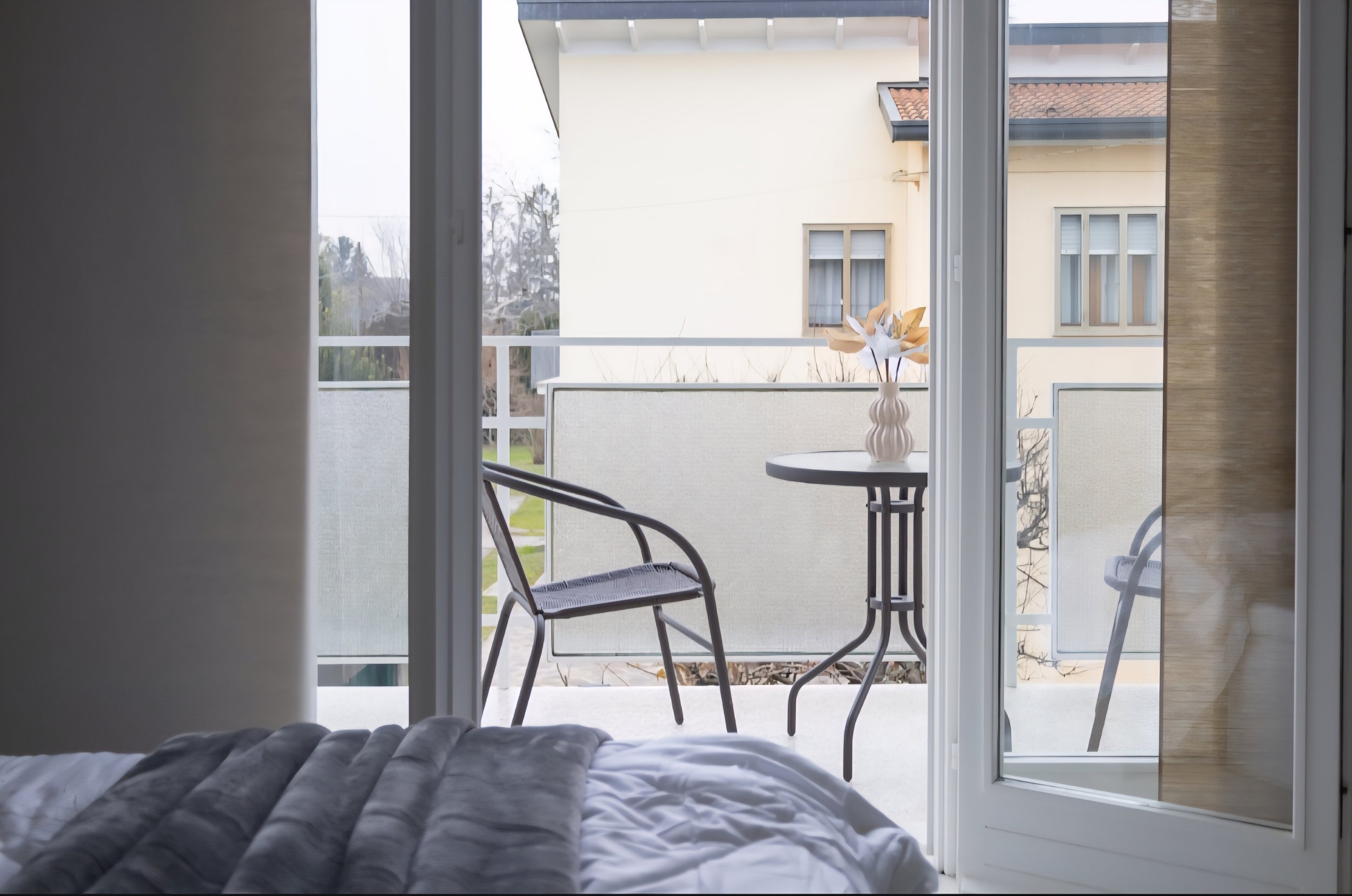 Vista di un balcone con sedia in metallo, tavolino con un vaso con fiori di carta, visto dall’interno di una stanza attraverso una porta a vetri