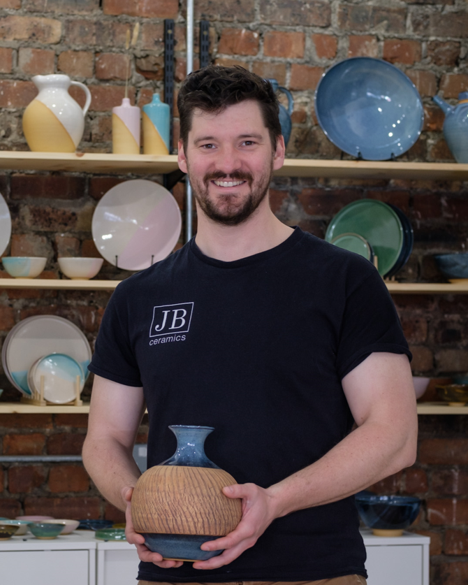 A man with dark hair and a beard smiling while holding a small ceramic vase in a pottery studio with shelves of ceramic dishes and a brick wall in the background.