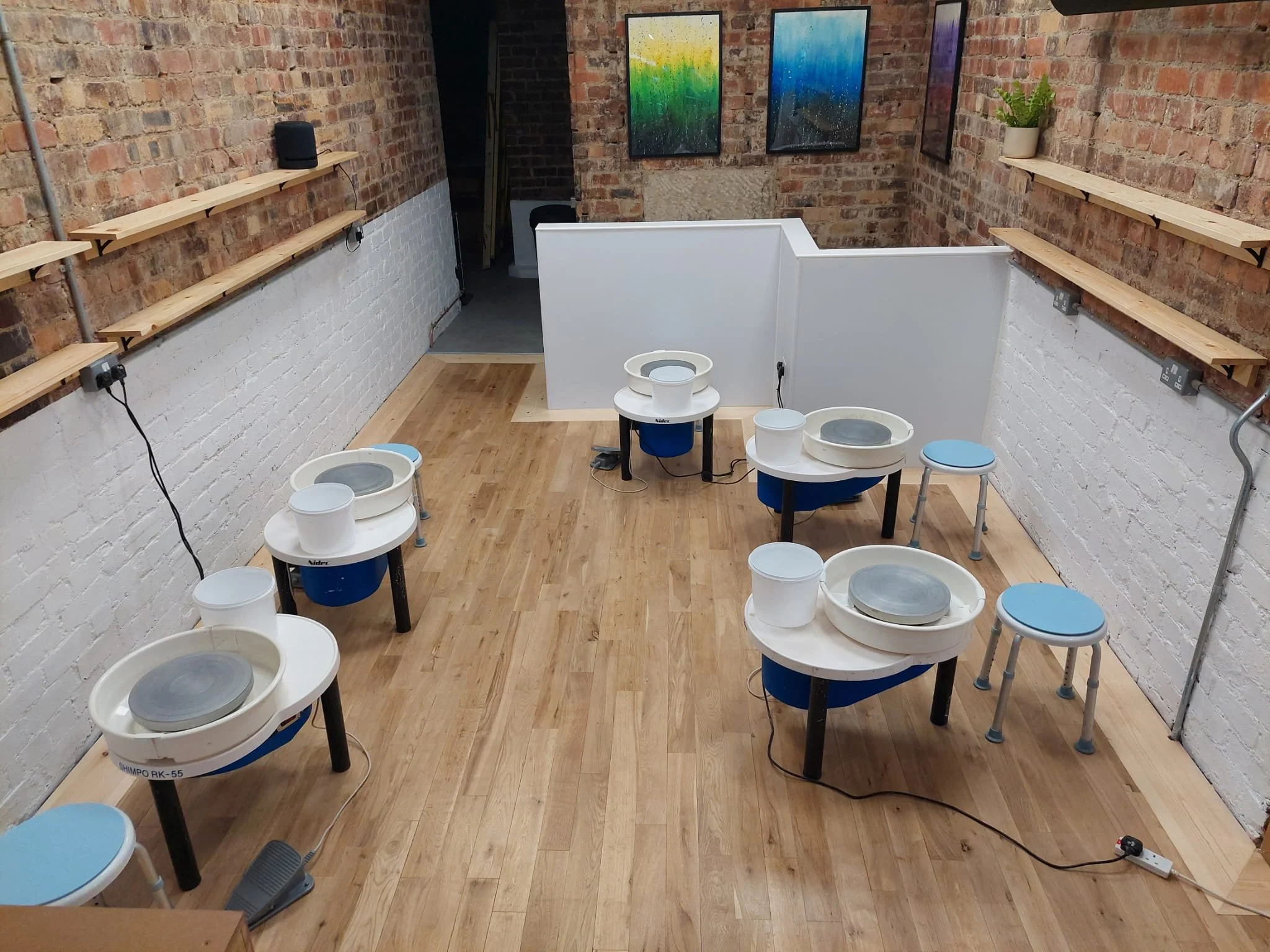 A pottery studio with six pottery wheels arranged in a semicircle on a wooden floor, with white and blue color schemes. The studio has brick and white painted brick walls, with colorful abstract paintings hanging on the brick wall. Two wooden shelves with plants and equipment are mounted on the brick walls, and a small stool with a blue cushion is near the wall. Cables and power outlets are visible.
