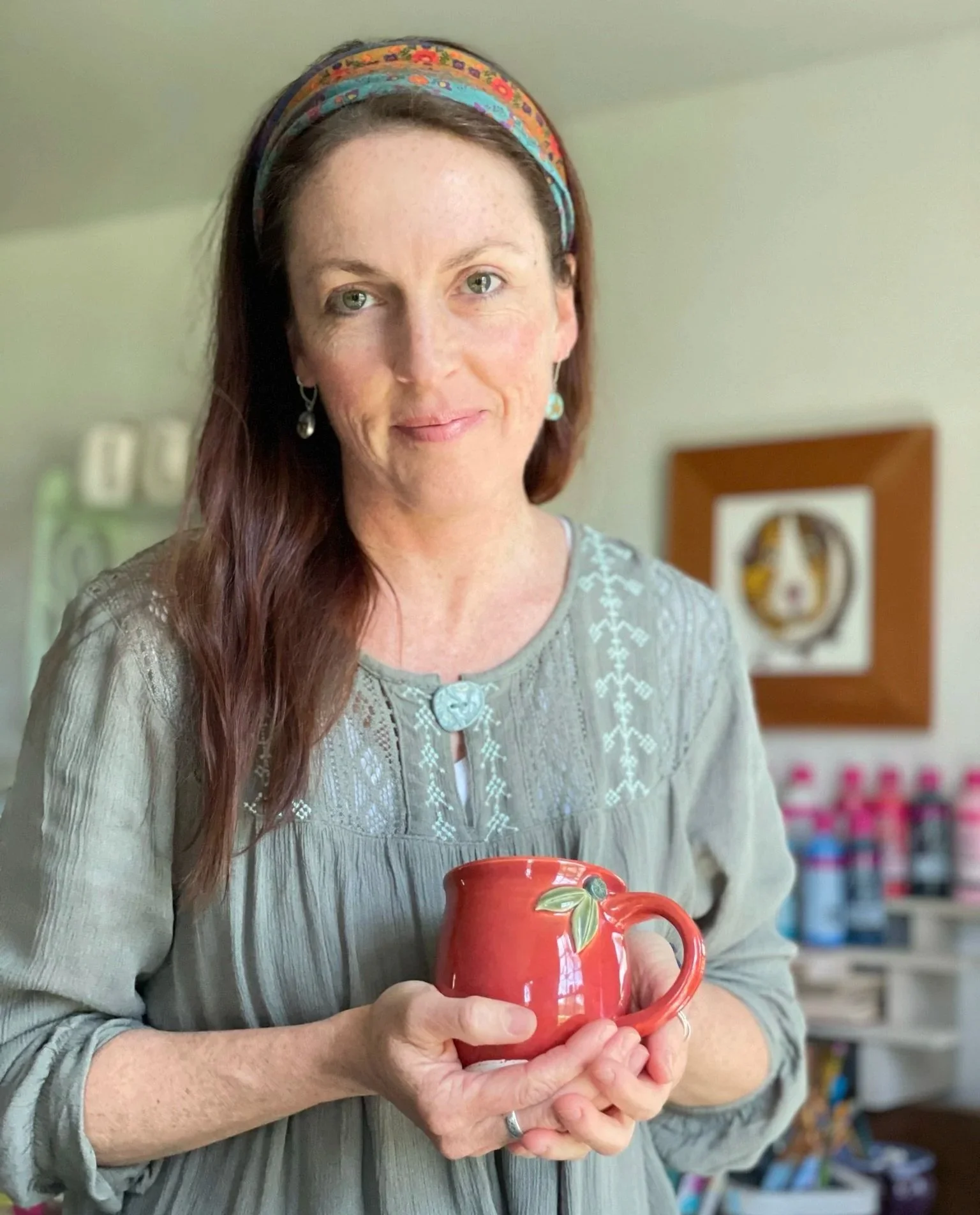 A woman with long brown hair, wearing a colorful headband and a gray embroidered blouse, holding a red mug with a leaf design, standing in a room with framed art and shelves in the background.