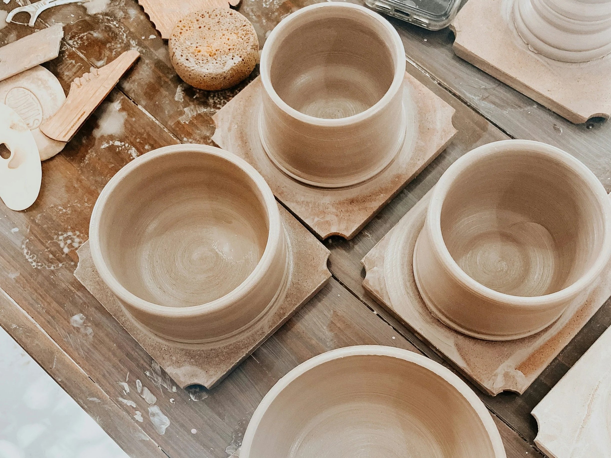 Four unfinished clay pots on a wooden worktable, with pottery tools and supplies nearby.