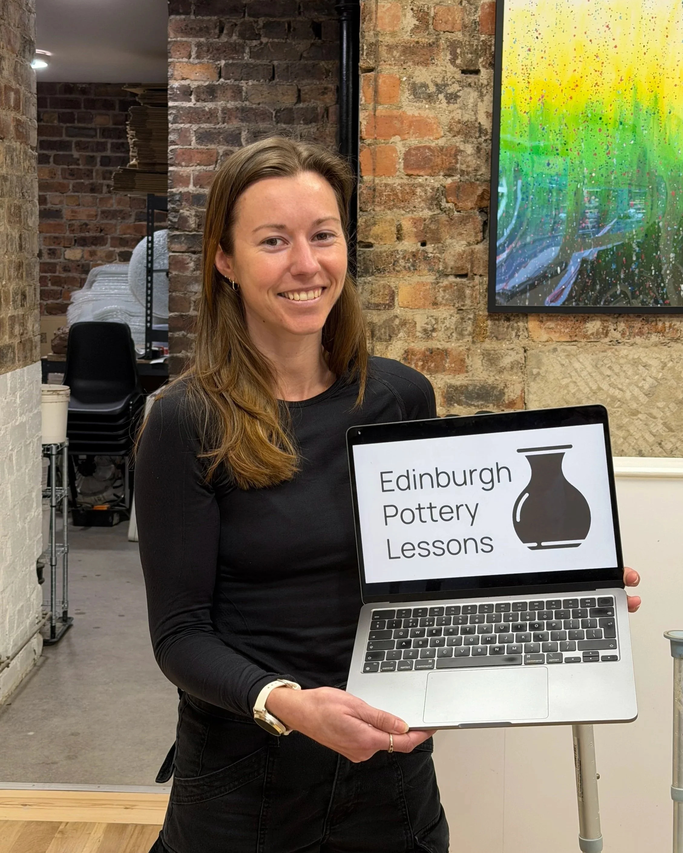 A woman with long brown hair smiling and holding a laptop displaying 'Edinburgh Pottery Lessons' with a pottery vase icon, inside a brick-walled room.
