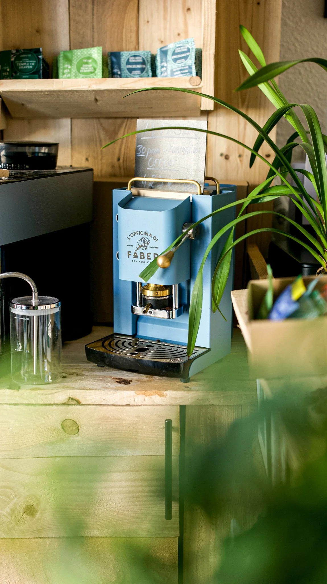 Une machine à espresso de couleur bleue sur une table en bois, avec une plante verte à droite et des sachets de thé ou café en arrière-plan sur une étagère en bois.