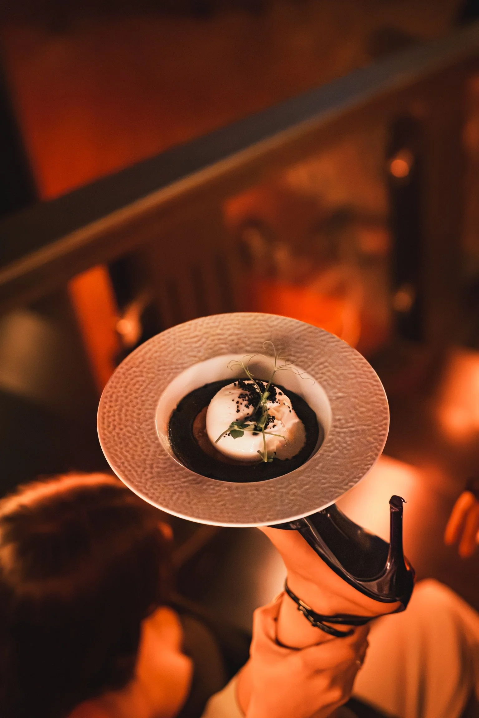 Une main tenant une cuillère à dessert avec un dessert mousseux ou crème, décoré de feuilles vertes et saupoudré de poudre noire, dans un bol blanc entouré d'une grande assiette en paille ou en matériau texturé. L'ambiance est chaleureuse avec un écl
