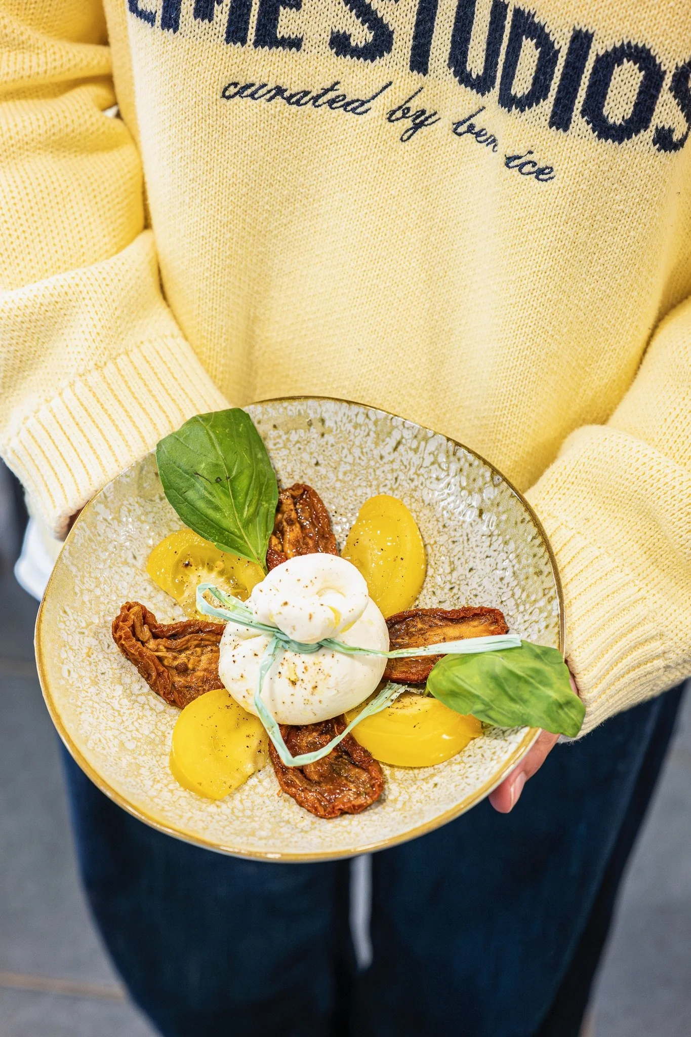 Une personne tient un bol contenant une salade avec des morceaux de tomate, du basilic, de la mozzarella et des feuilles de salade vertes. La personne porte un sweat-shirt jaune avec du texte en anglais.
