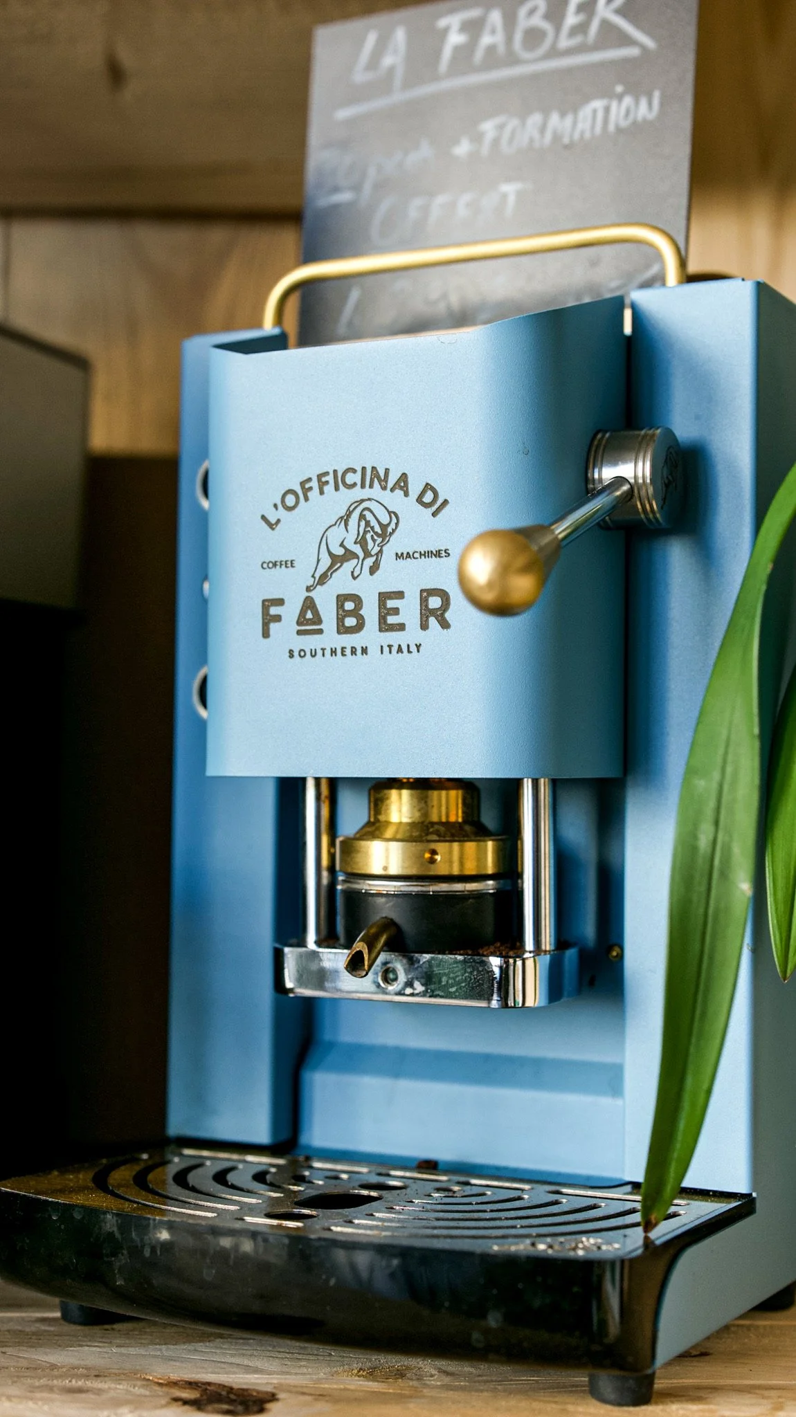Une machine à café de couleur bleu clair, portant le logo « L'Officina Di Faber », située sur un plan de travail en bois. On peut voir une tige en métal avec une boule dorée, qui est probablement une pièce de la machine. En arrière-plan, un panneau n