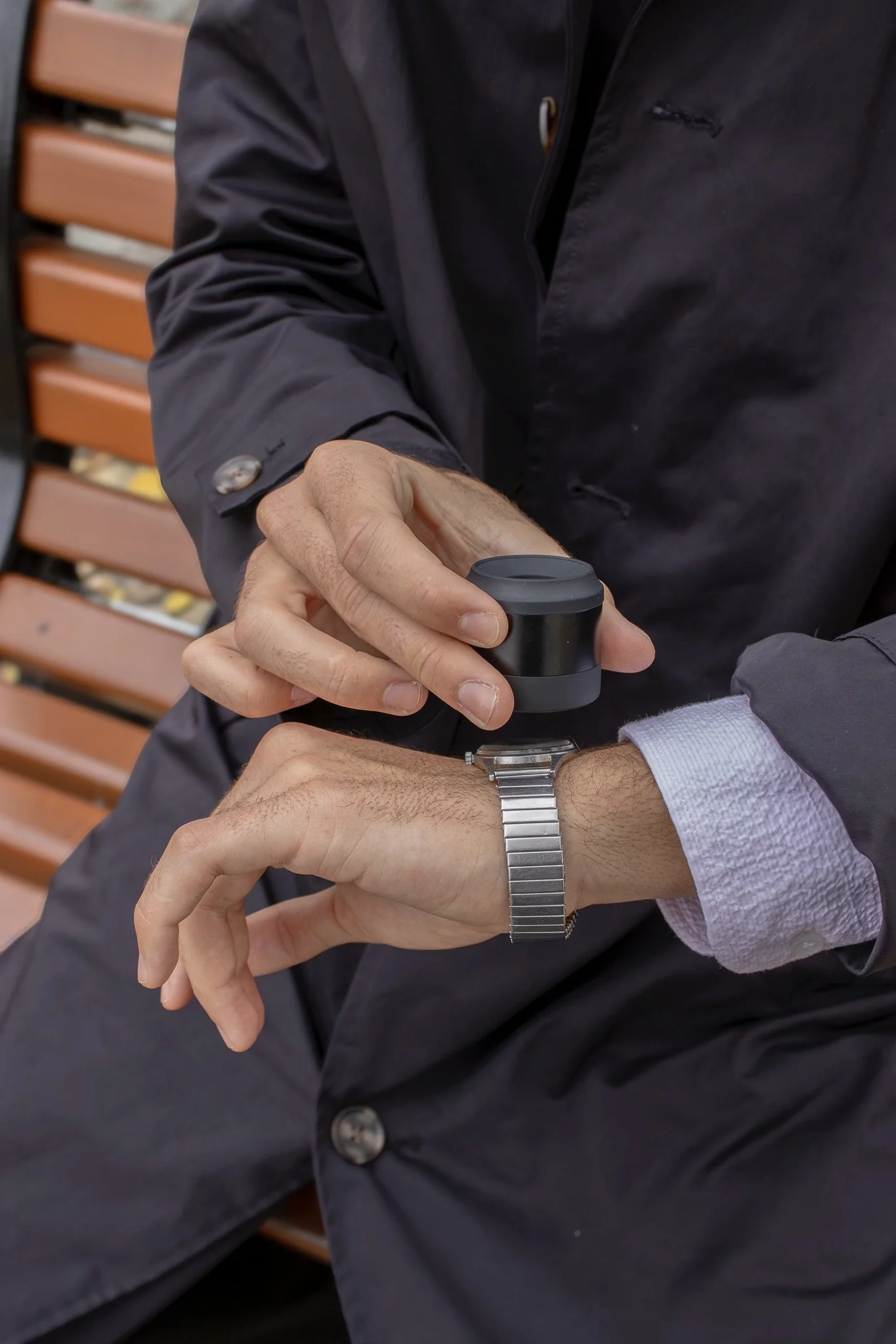 Une personne tenant une montre ou un objet noir rond, habillée d'une veste noire, avec une autre personne dont l'avant-bras est visible portant une montre en métal, dans un endroit avec un banc en bois en arrière-plan.