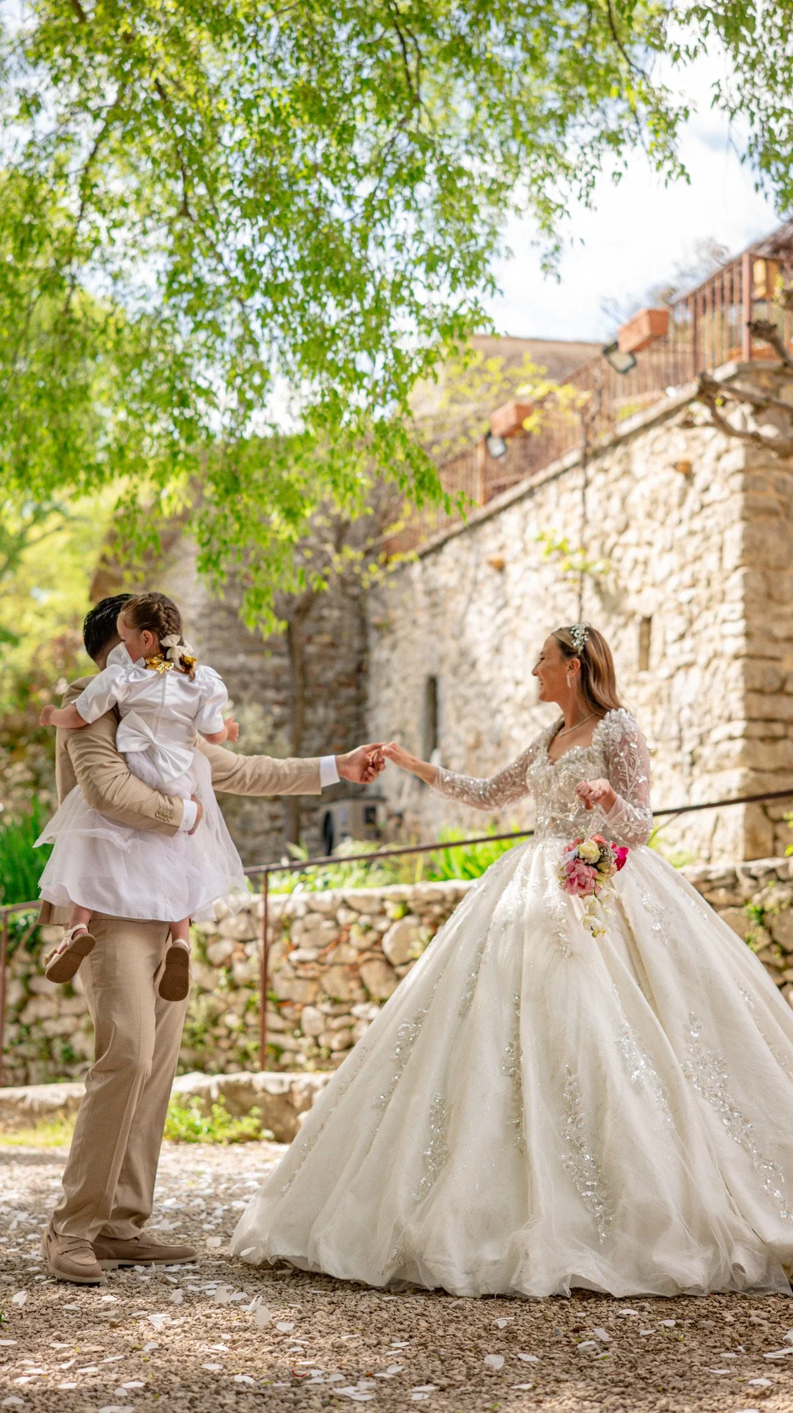 Une mariée en robe blanche avec une jupe volumineuse et des détails en dentelle, échangeant un geste avec une femme portant un pantalon beige et une veste, dans un cadre extérieur avec des arbres et un mur en pierre en arrière-plan.