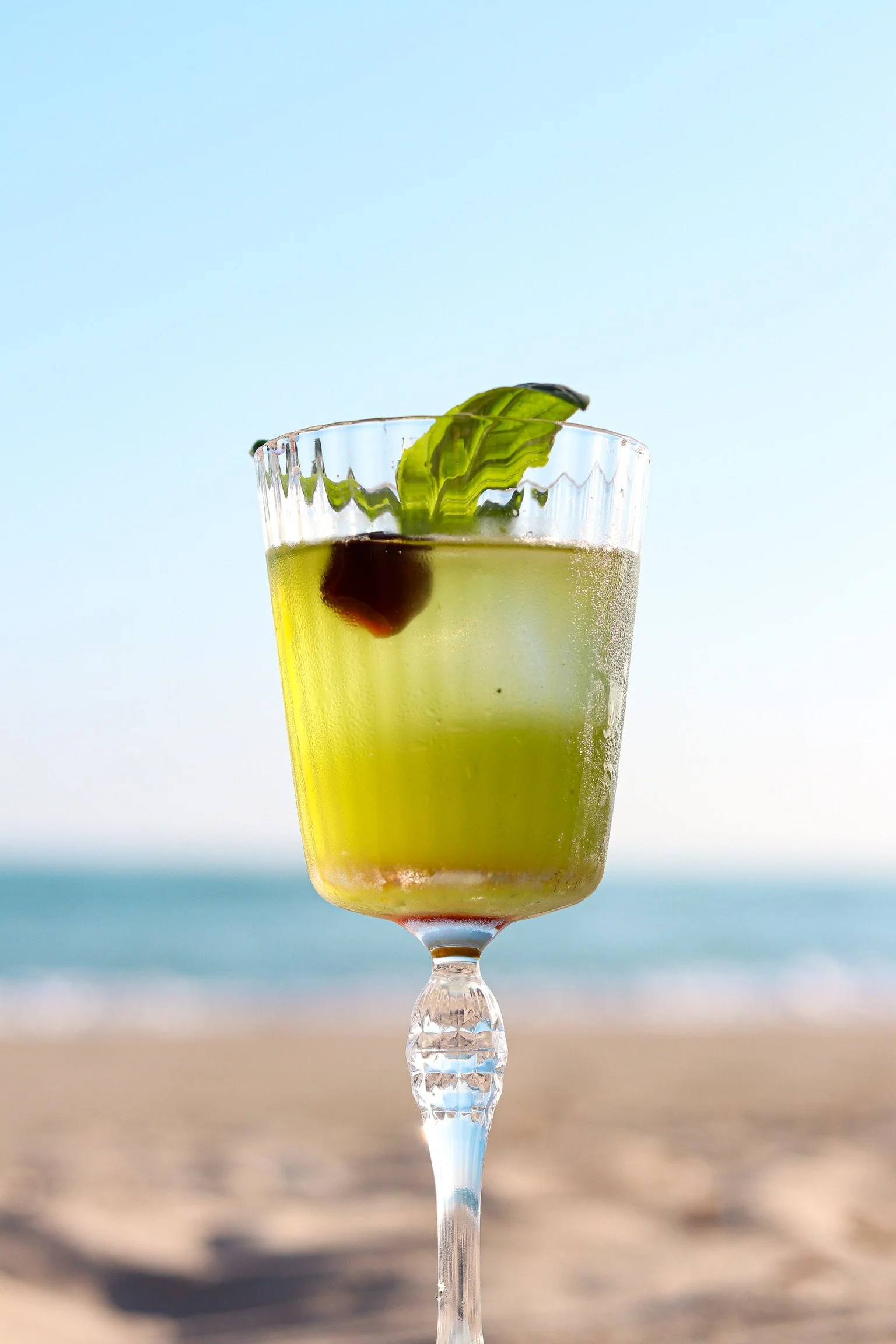 Un verre à cocktail rempli d'une boisson jaune, décorée avec une olive et une feuille de menthe, sur un fond de plage et de mer.