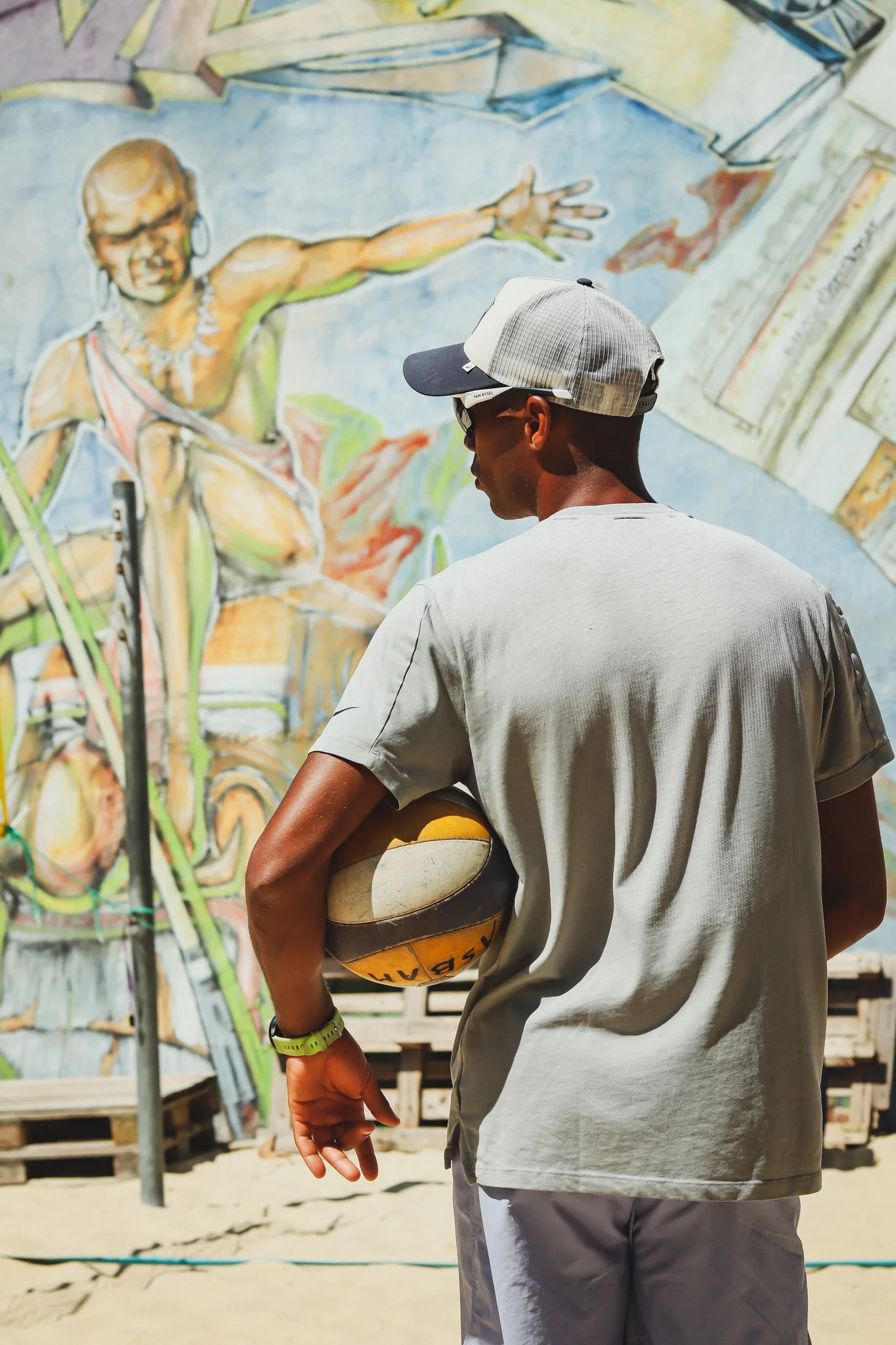 Un jeune homme portant une casquette, un t-shirt gris, un bracelet vert, tenant un ballon de basketball, se tient devant une fresque murale représentant une femme qui pointe du doigt.