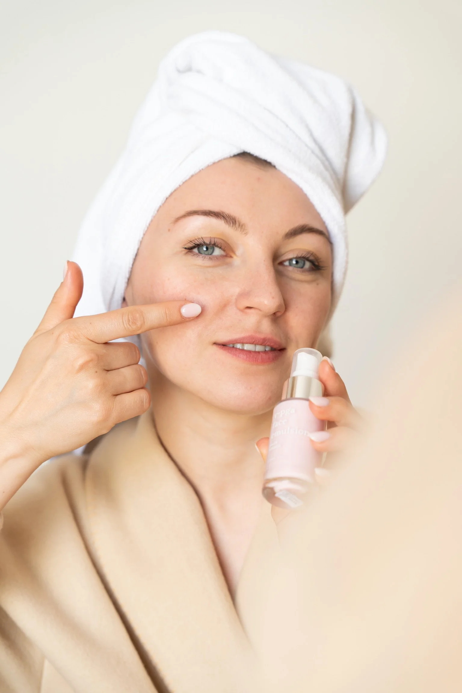 Une femme avec un turban blanc appliquant une crème ou un produit de soin facial, portant un manteau beige.