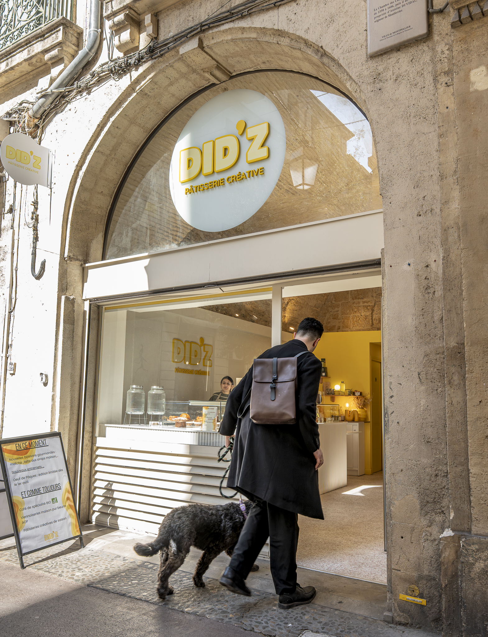 Une personne portant un manteau noir, avec un sac à dos marron, marche avec un chien noir à l'entrée d'une pâtisserie créative nommée Did'z. Le magasin a une devanture en verre, avec un panneau d'affichage à gauche.