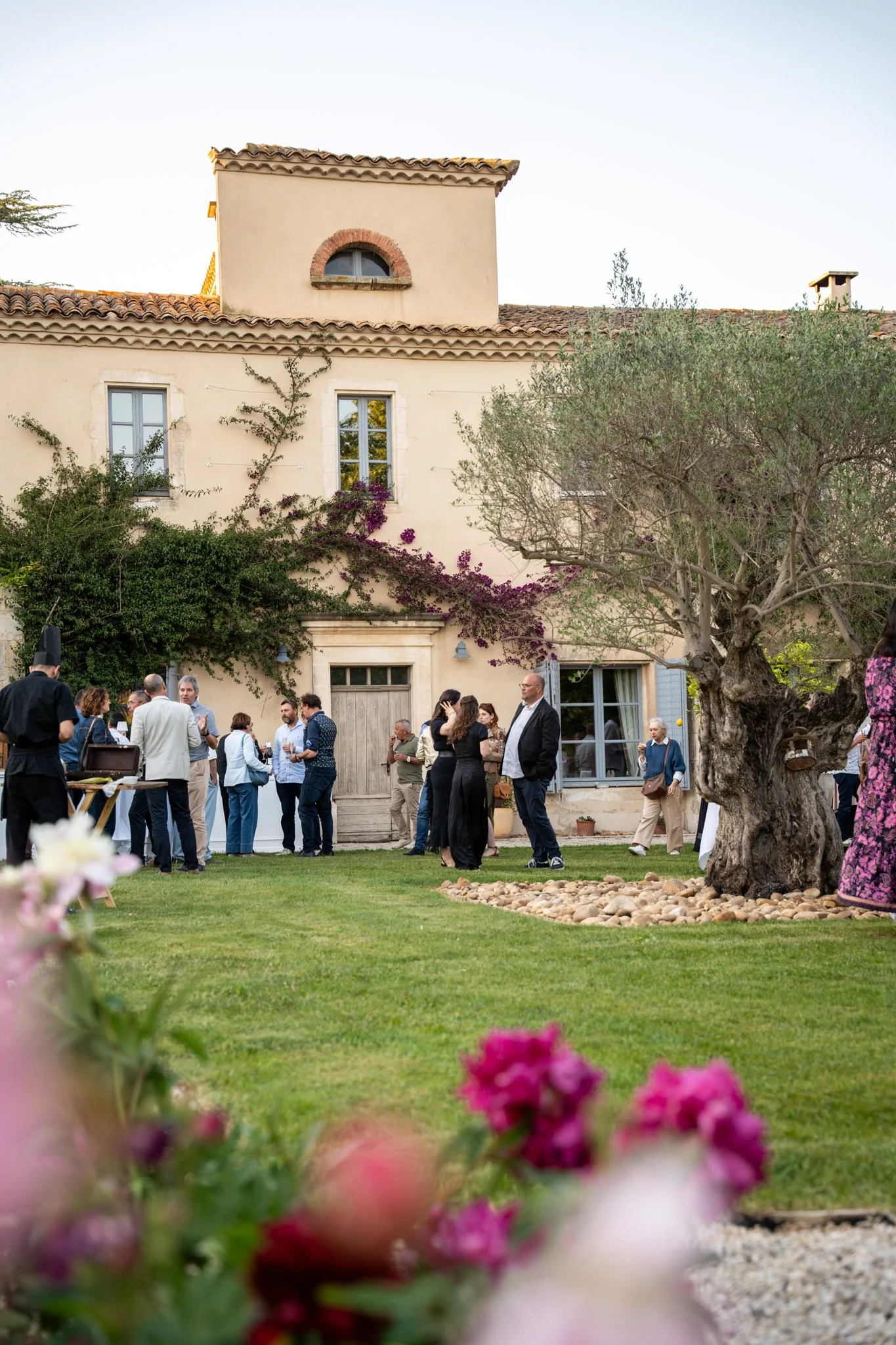 Groupe de personnes en costume lors d'une réception en extérieur devant une maison en pierre avec un jardin et des arbres.