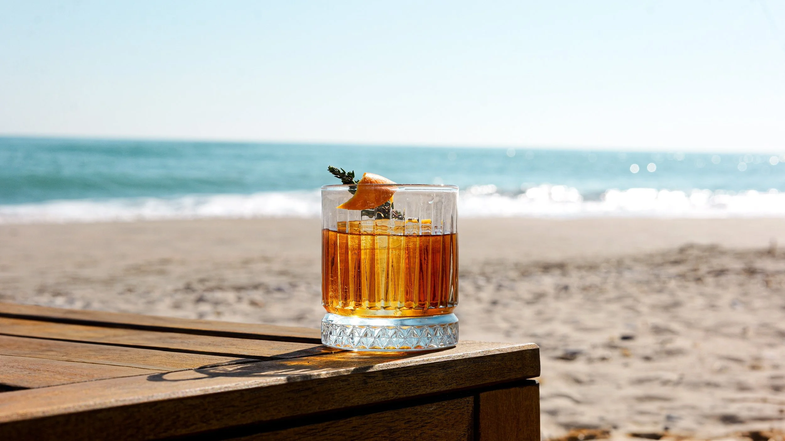 Un verre de cocktail brun-orange avec une tranche d'orange et une branche de thym, posé sur une table en bois à la plage avec la mer et le ciel bleu en arrière-plan.