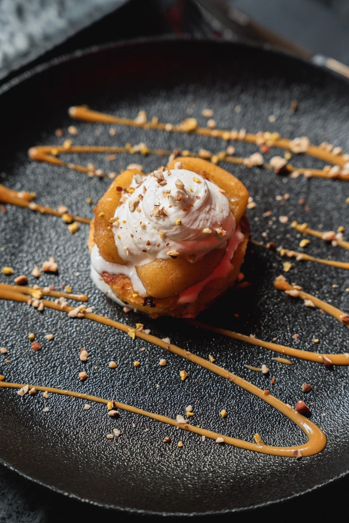 Un dessert de pâtisserie avec une couche de chantilly, des morceaux de bananes et des noisettes, présenté avec un dessin de caramel sur une plaque noire.