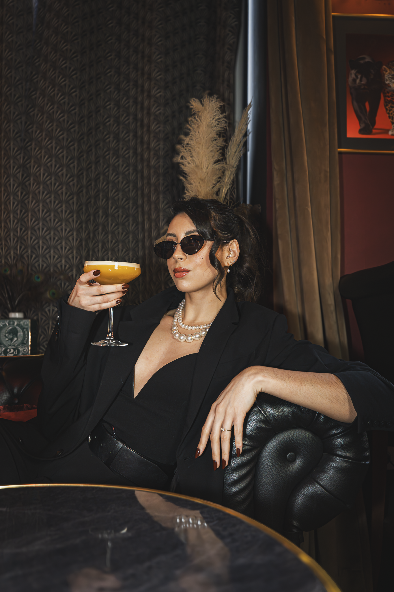 Femme élégante portant des lunettes de soleil, un collier de perles, vêtue de noir, assise dans un fauteuil dans un environnement sophistiqué.