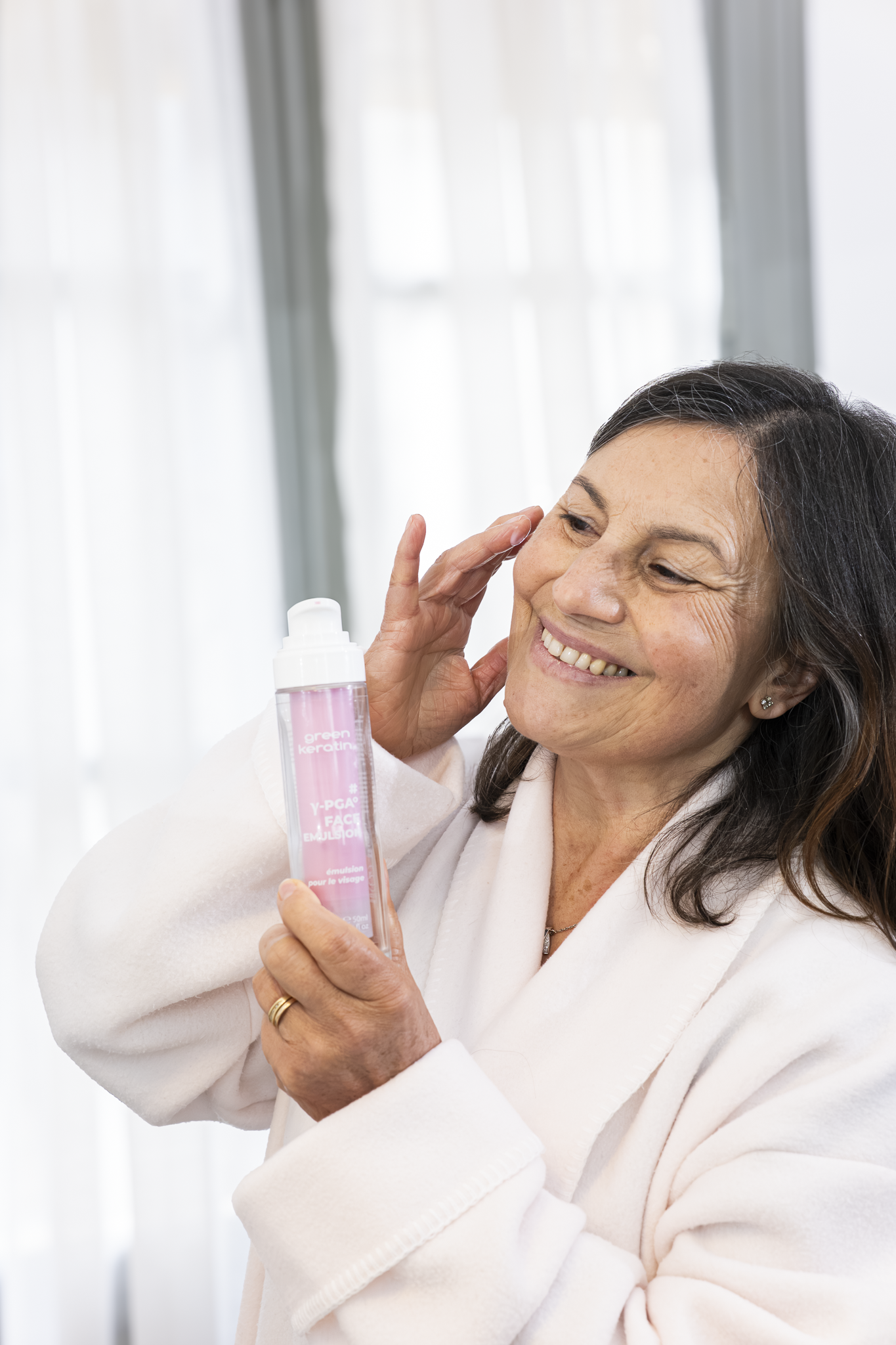 Une femme âgée souriante, portant une robe blanche et un gilet blanc, tient un flacon de produit de soin crème pour le visage, dans une pièce lumineuse avec des rideaux blancs.