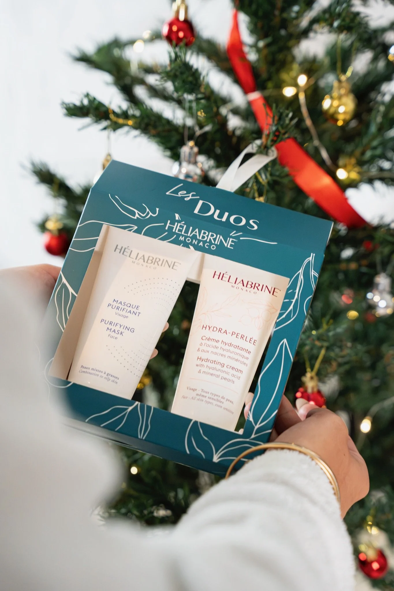Une main tenant un coffret cadeau contenant des produits de soins pour la peau, avec un sapin de Noël décoré en arrière-plan.