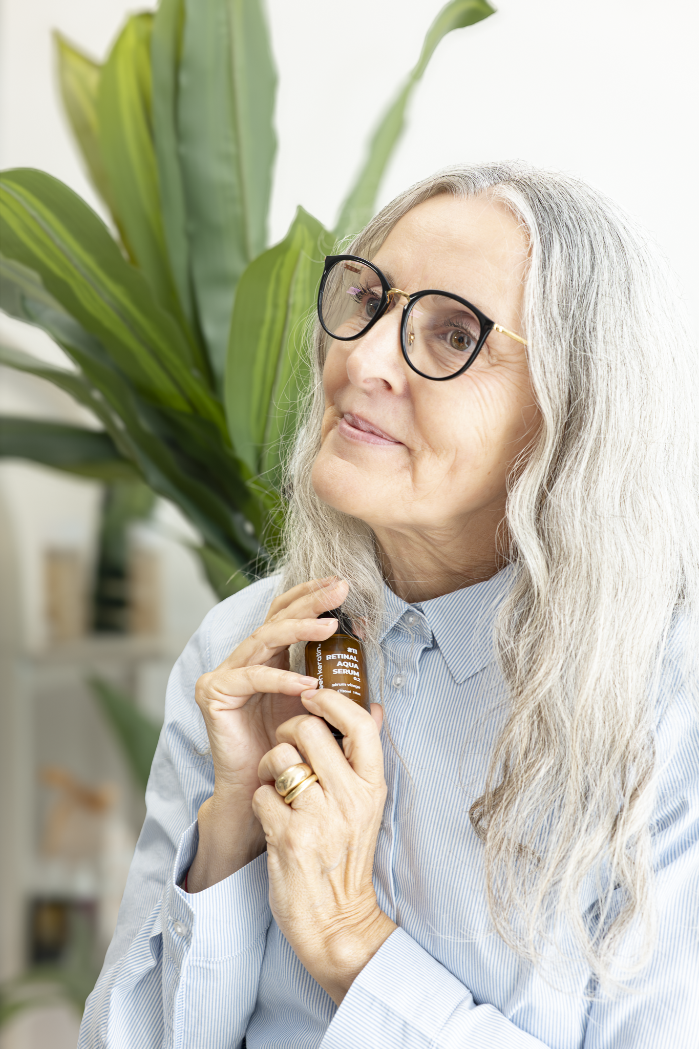 Une femme âgée aux cheveux gris, portant des lunettes à monture noire et or, tient un flacon de sérum visage dans ses mains. Elle est habillée d'une chemise à rayures bleues et blanches, et se trouve à côté d'un grand plant vert. L'arrière-plan est f