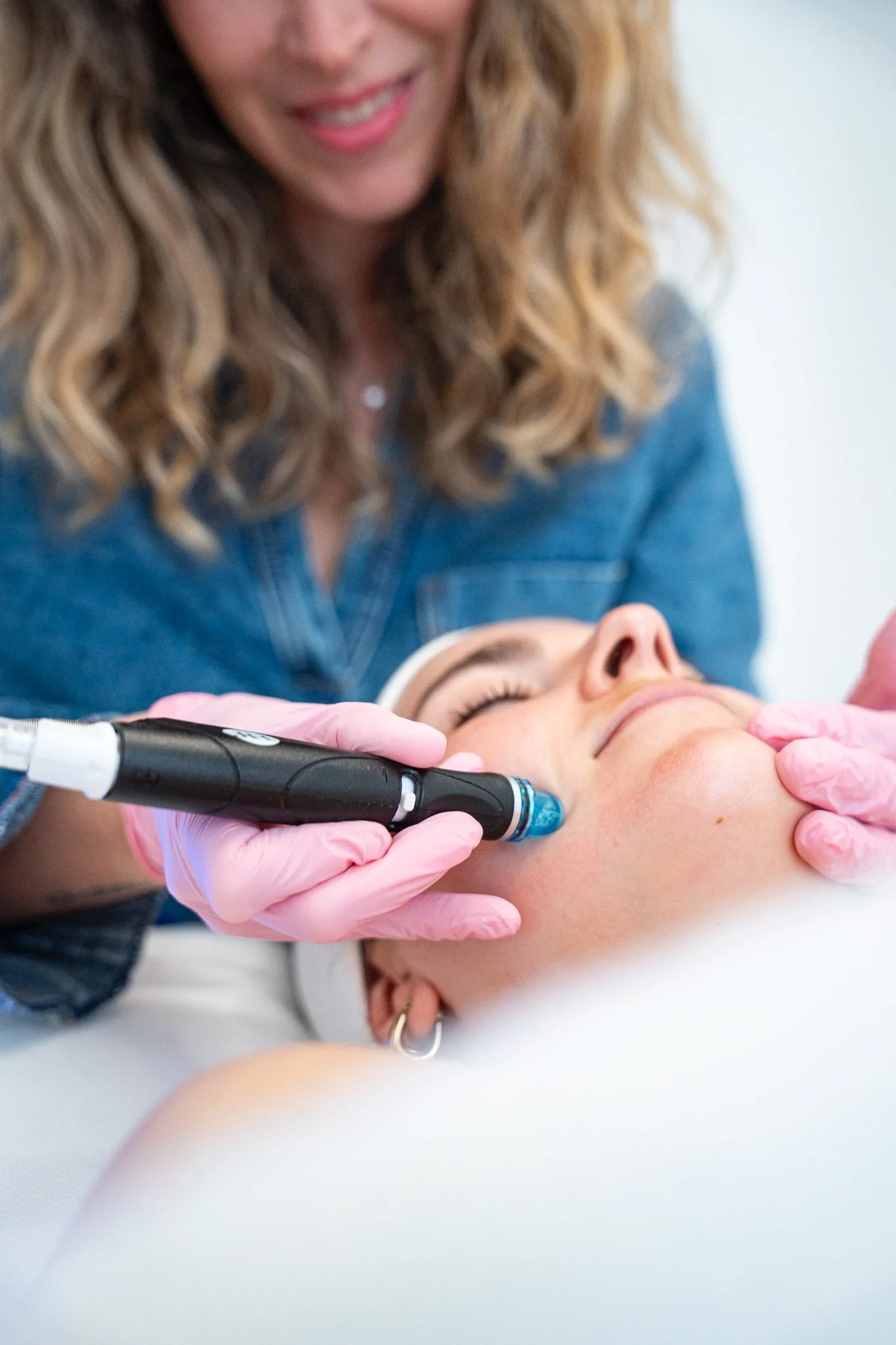 Une femme effectuant une injection au visage d'une autre personne allongée, avec des mains gantées. La femme sourit, portant une chemise en denim et a de longs cheveux bouclés. La personne recevant l'injection a les yeux fermés.