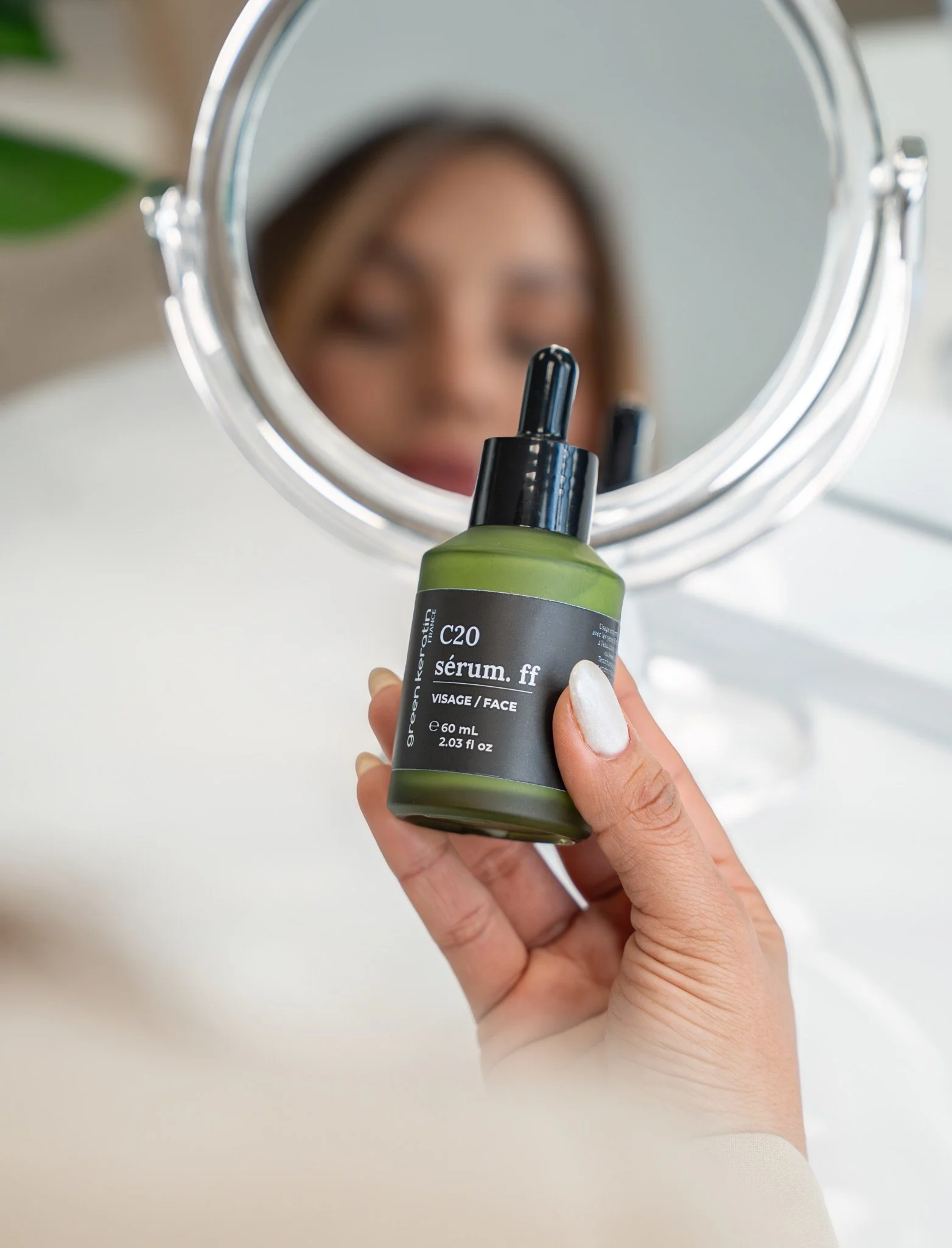 Une main tenant un flacon de sérum facial devant un miroir, avec le reflet d'une personne floue en arrière-plan.