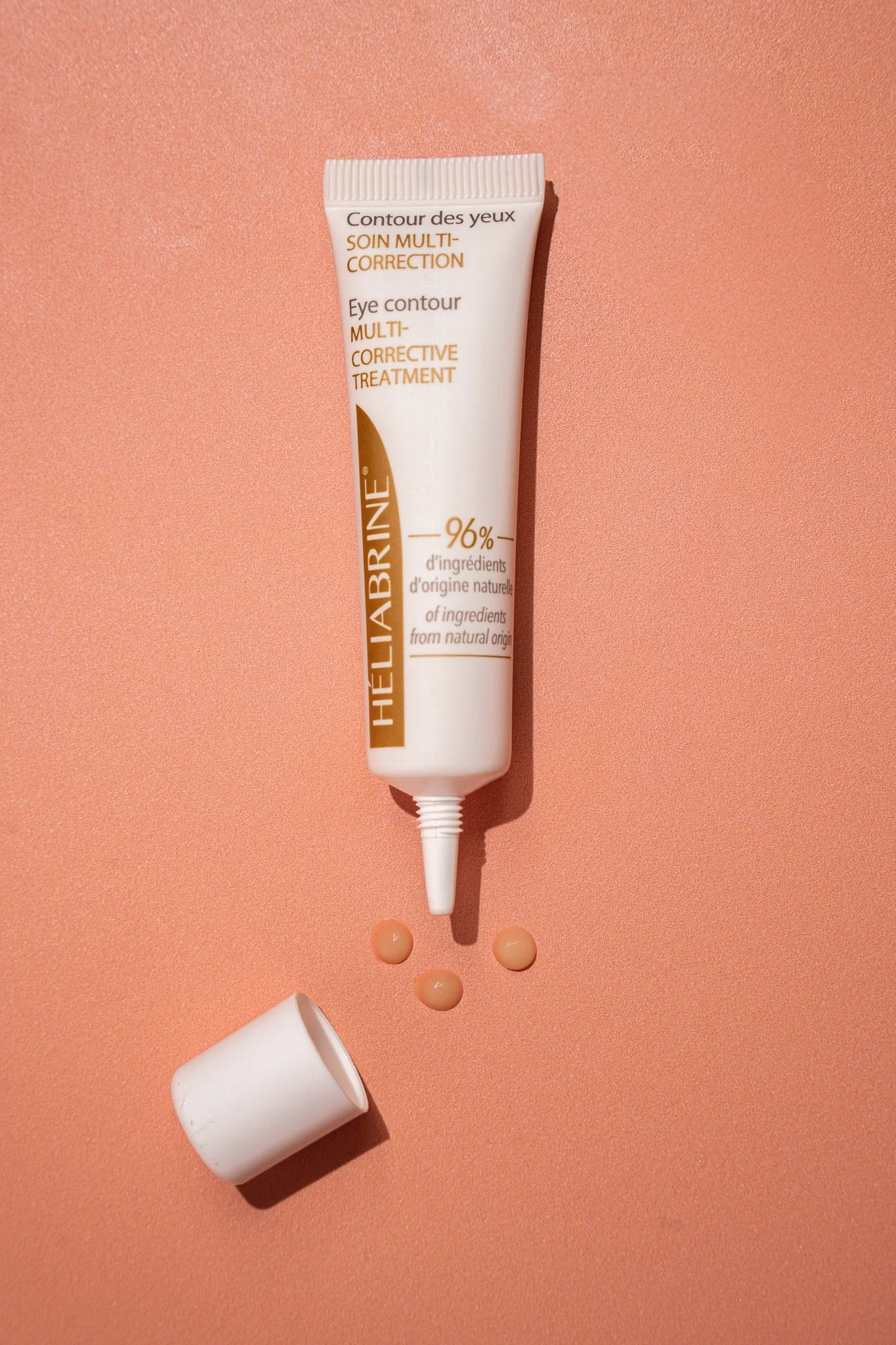 Tube de crème pour le contour des yeux avec des gouttes de crème et un bouchon blanc posé à côté, sur un fond couleur pêche.