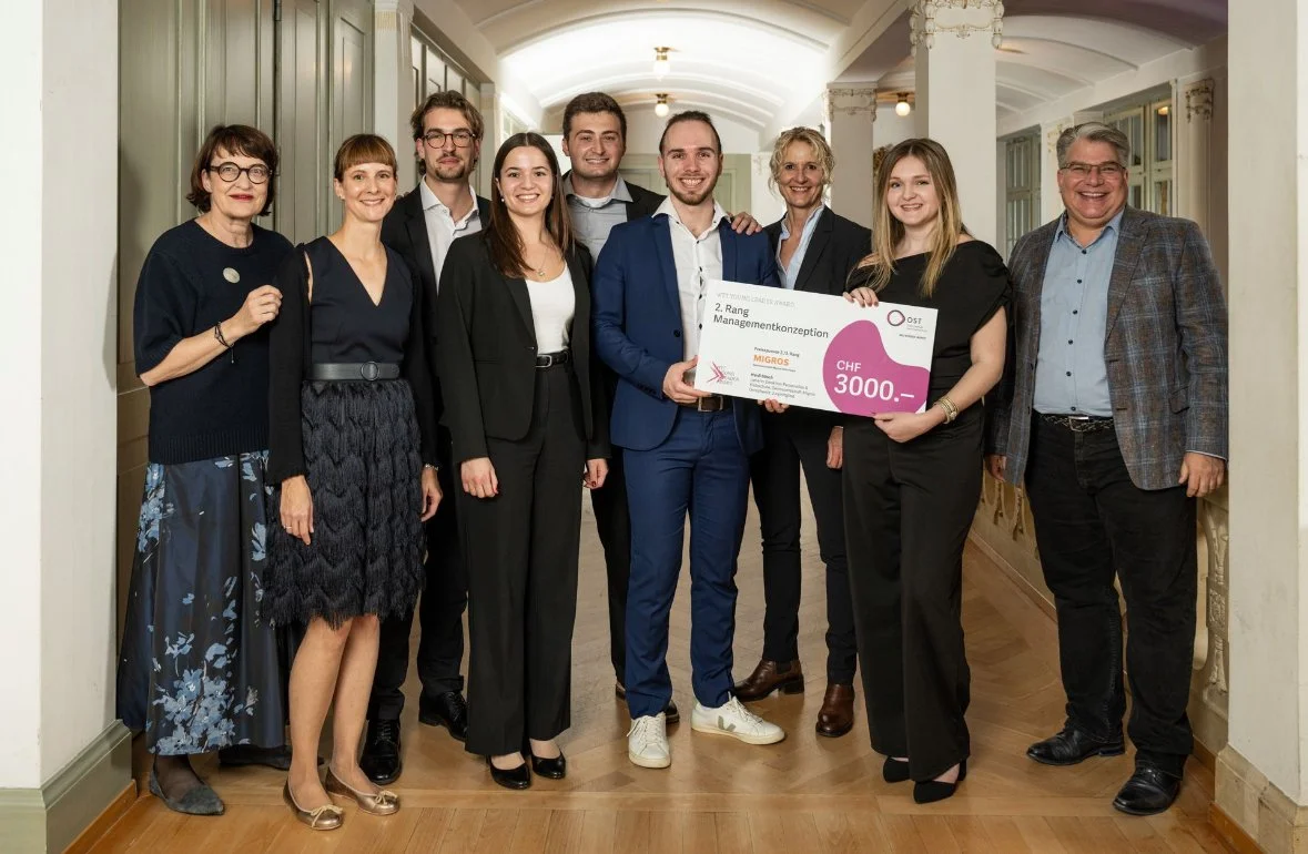 Group of nine people standing in a hallway, celebrating an award or prize, with one person holding a large check for 3000 Swiss Francs.