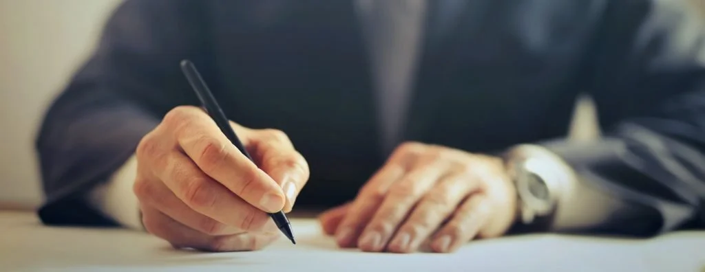 A person in a suit is writing on paper with a black pen, sitting at a desk.